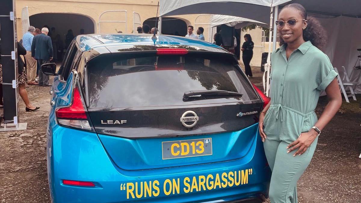 Biologist Shamika Spencer poses next to a blue Nissan Leaf car on which the sentence "Runs on Sargassum" is printed. 