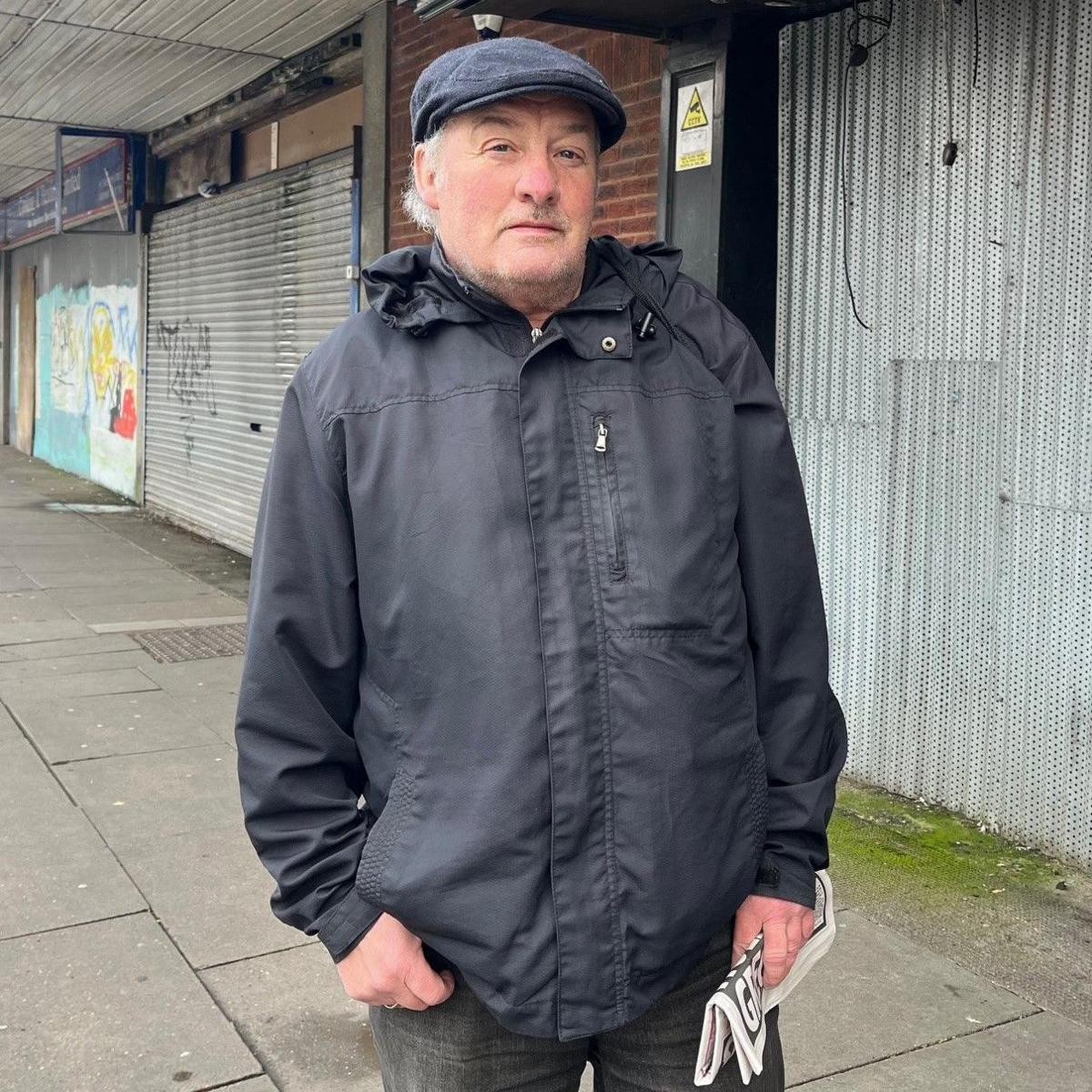 John Holmes carries a rolled-up copy of a newspaper as he stands in Freeman Street, Grimsby, with boarded-up shops behind him. He is wearing a grey jacket and a blue flat cap