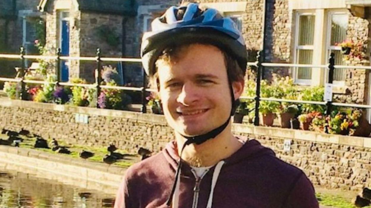 Max McGhee wearing a hoody and a bike helmet. He is stood next to a river and there are stone houses behind him. 