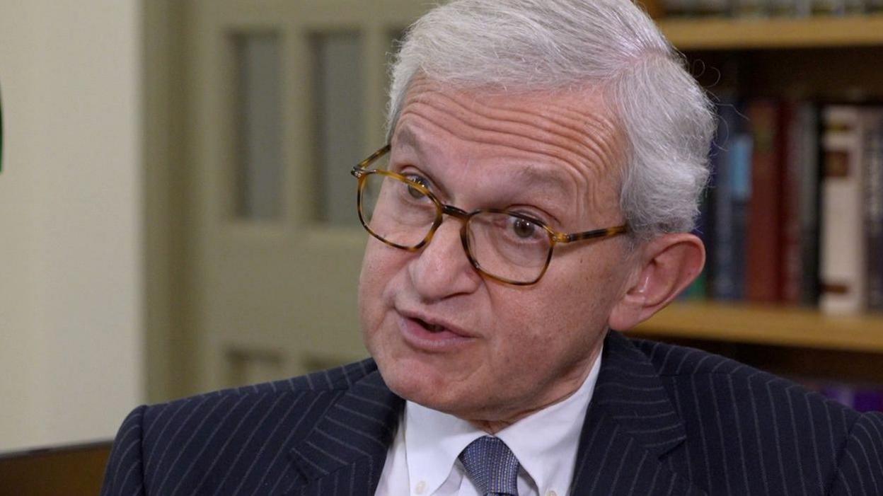 Sir Geoffrey Vos has grey short hair and is wearing glasses. He is dressed in a pinstripe navy suit and is wearing a white shirt and tie. There is a bookshelf behind him