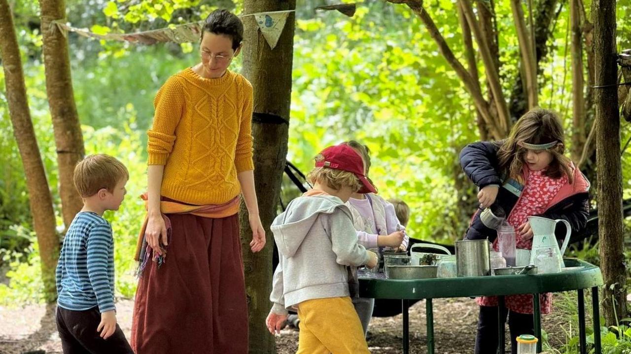 A woman in a yellow jumper and red skirt plays with children in a forest. There is a table covered with lots of cooking utensils. 