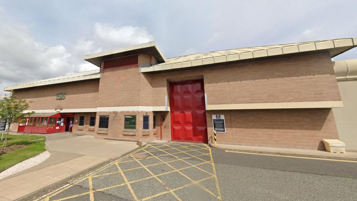 Front of a prison. It is a large brick wall with a large red door for vans and some windows.