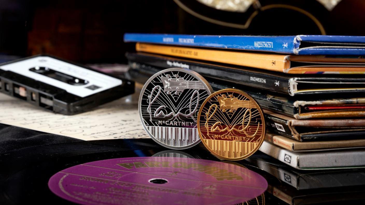 A silver and gold coin upright, side by side, with Paul McCartney writing on, leaning next to a tape and books