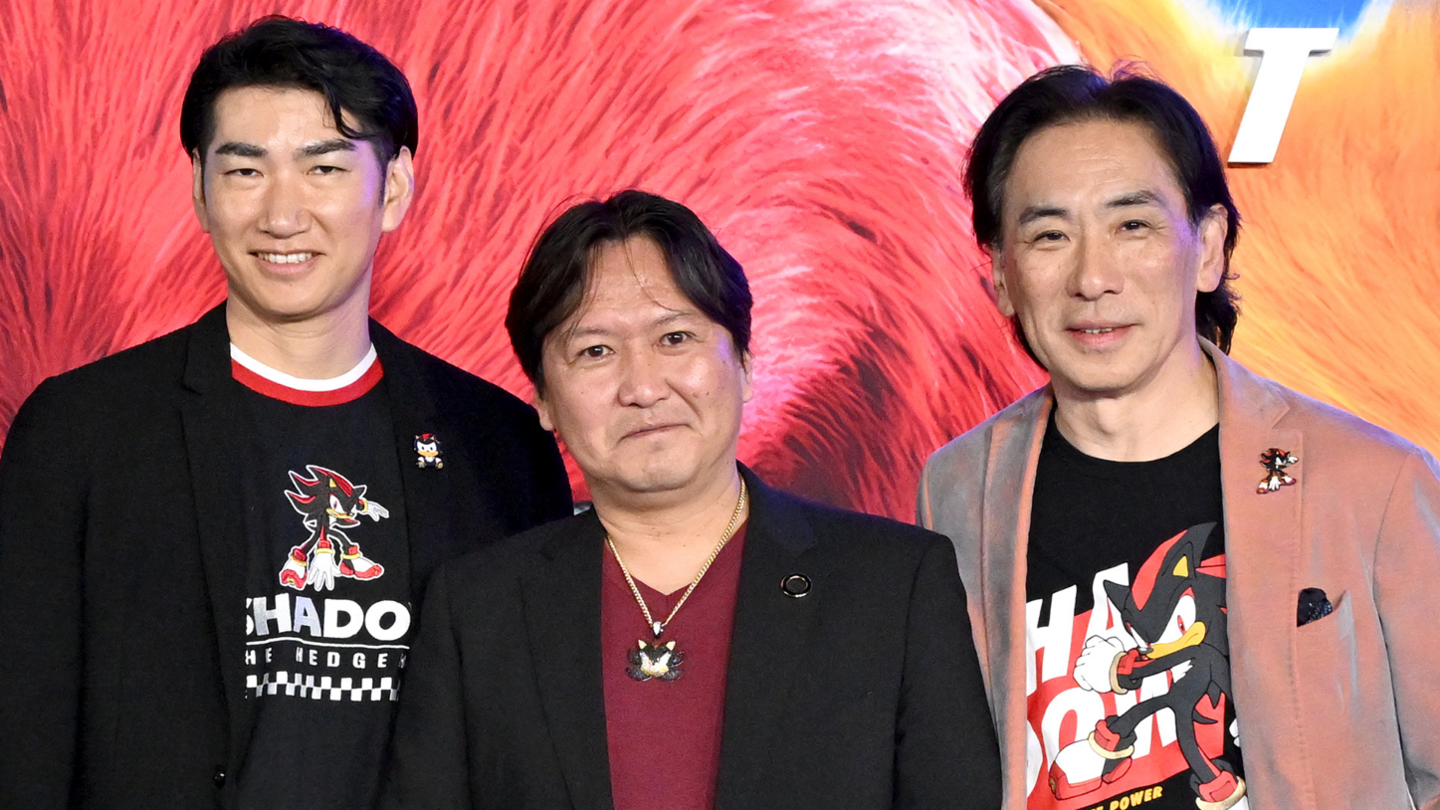 Three Japanese men stand together smiling for a photograph at a film premiere. Behind them is a backdrop featuring characters in the Sonic movie. The three men are each wearing an item of clothing featuring Shadow the Hedgehog, who features in the film. Satomi and Utsumi have Shadow on a t-shirt as well as lapel badges, while Iizuka has a Shadow necklace.