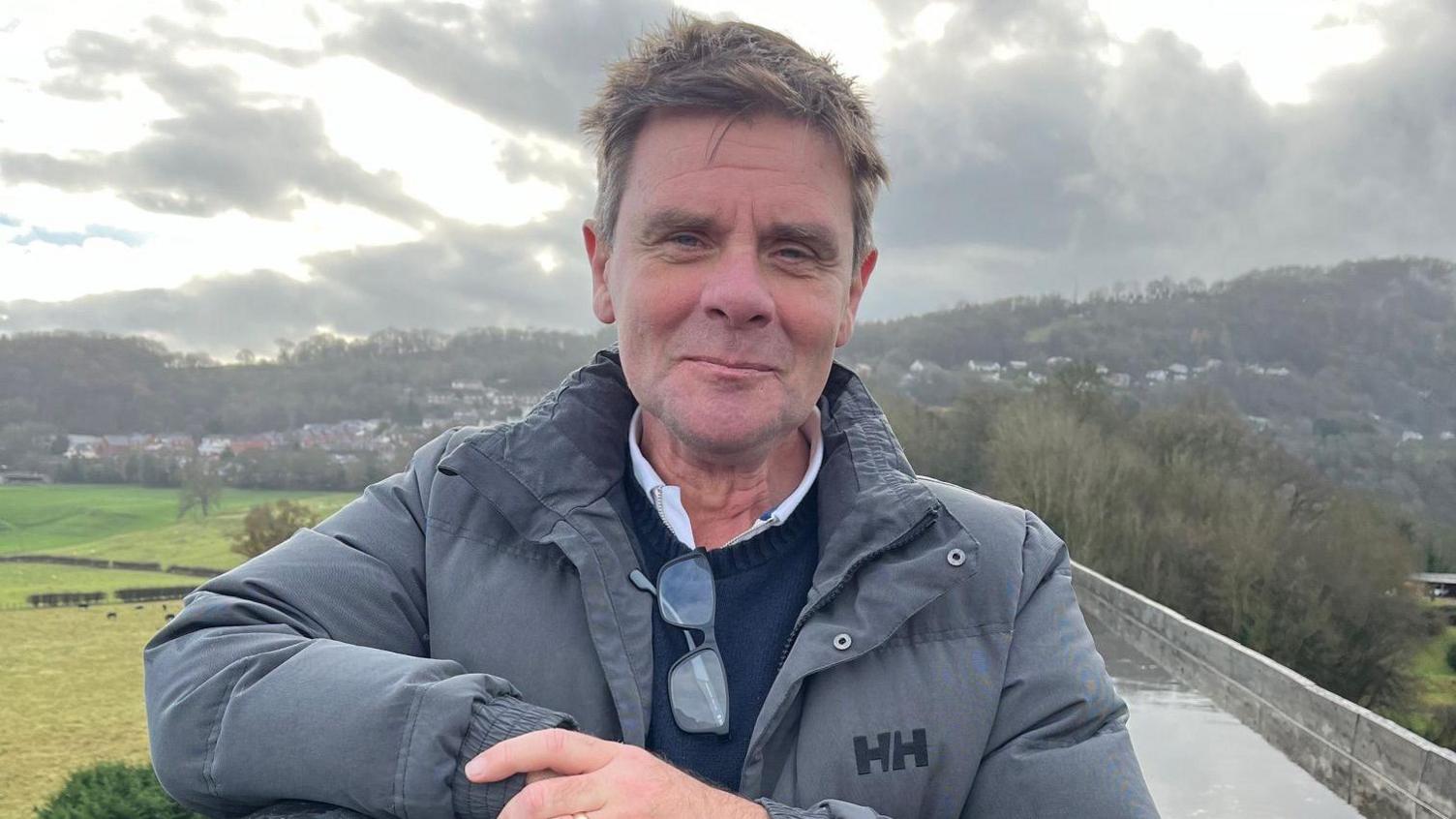 A man with brown hair and a slight smile is stood outside on what appears to be a bridge with scenery behind him - a hill on the right and fields on the left. He is wearing a grey thick jacket with a Helly Hanson logo on it. He wears a white shirt, a blue jumper over it and his glasses are hanging on the collar of his jumper.