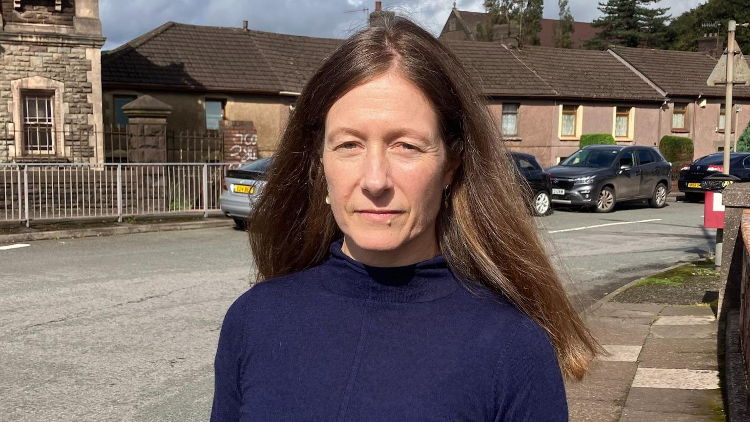 Louise Miskell wearing a navy blue jumper with long brown hair. She is looking at the camera while standing in front of houses