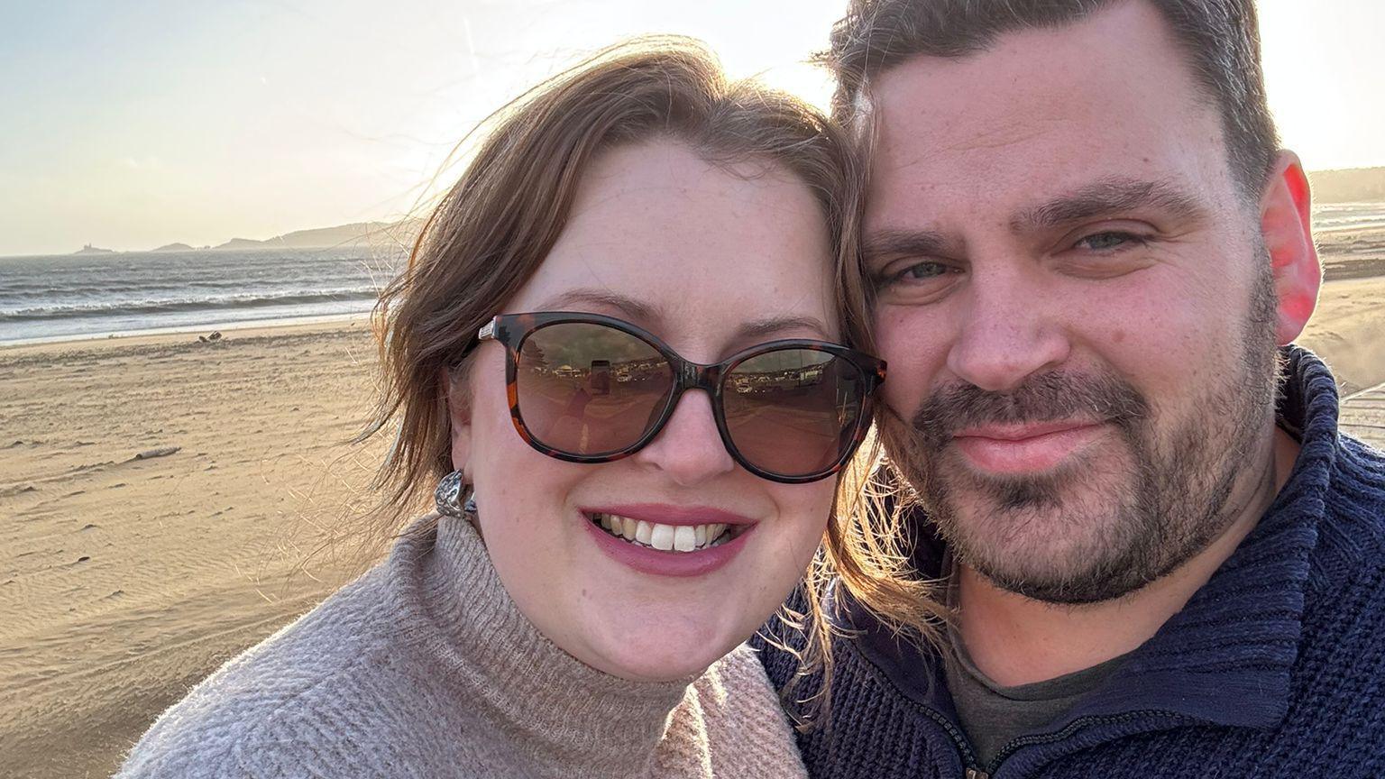 Jen and her husband are on a beach, standing close together looking at the camera. She is wearing sunglasses and a beige knitted polo-neck jumper. Chris is wearing a navy knitted sweater.