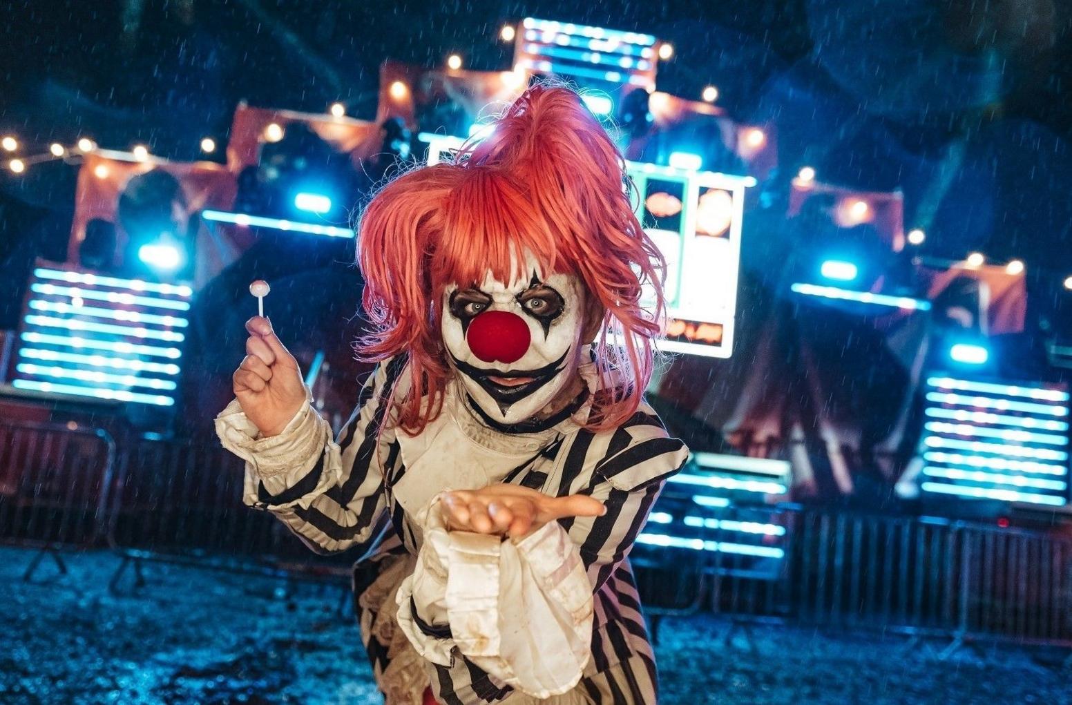 An actor dressed as a scary clown with a red wig blows a kiss at the camera while behind them an illuminated fairground ride is visible