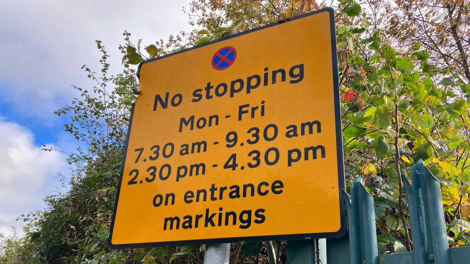 A yellow traffic sign outside a school in Leeds reads NO STOPPING Mon - Fri 7.30am - 9.30am 2.30pm - 4.30pm on entrance markings 