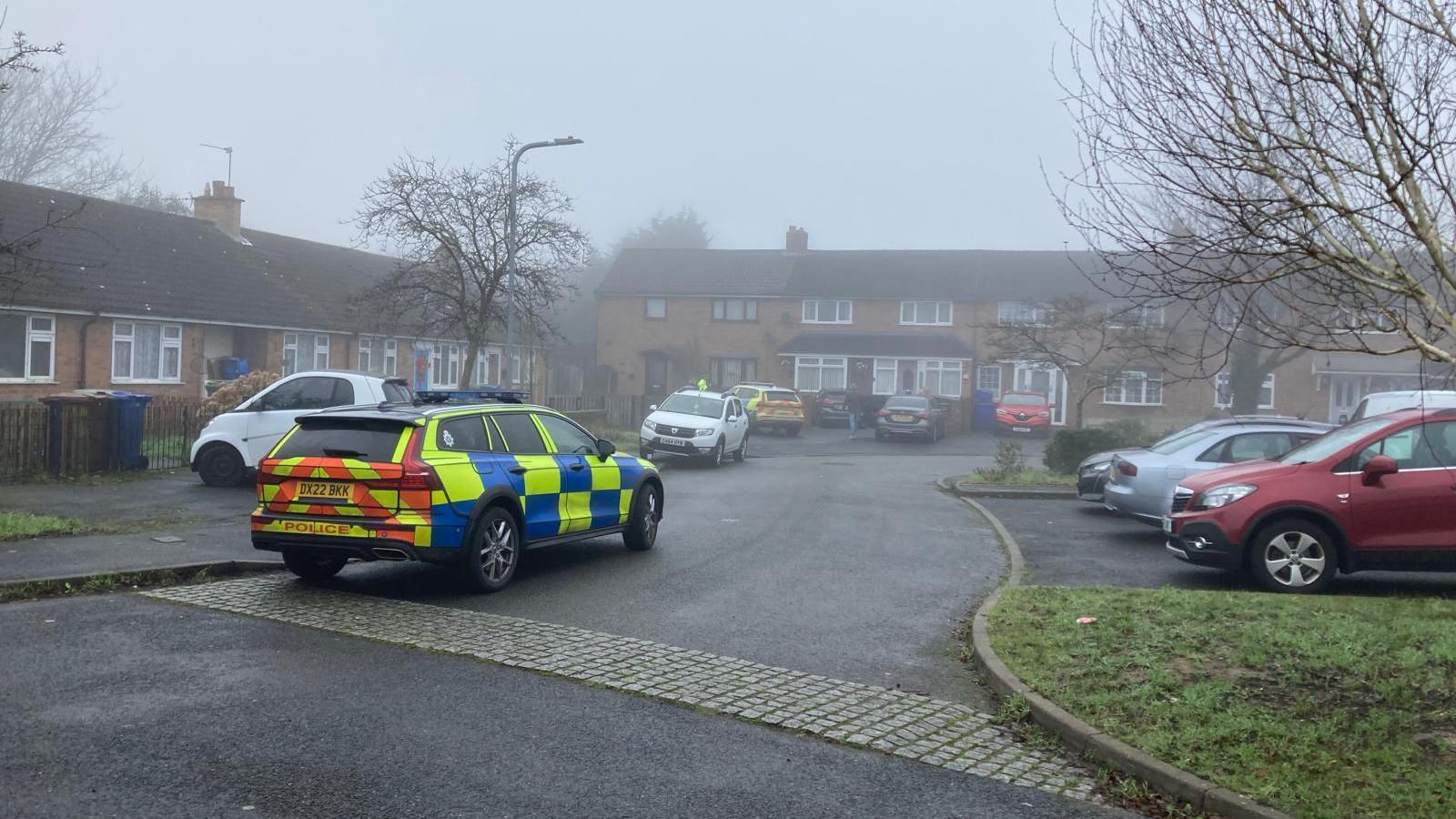 The corner of a residential street on a misty day with a police car parked at the side of the road on the left and another parked in a house's driveway in the distance. A number of other non-emergency vehicles are parked in driveways and at the side of the street.