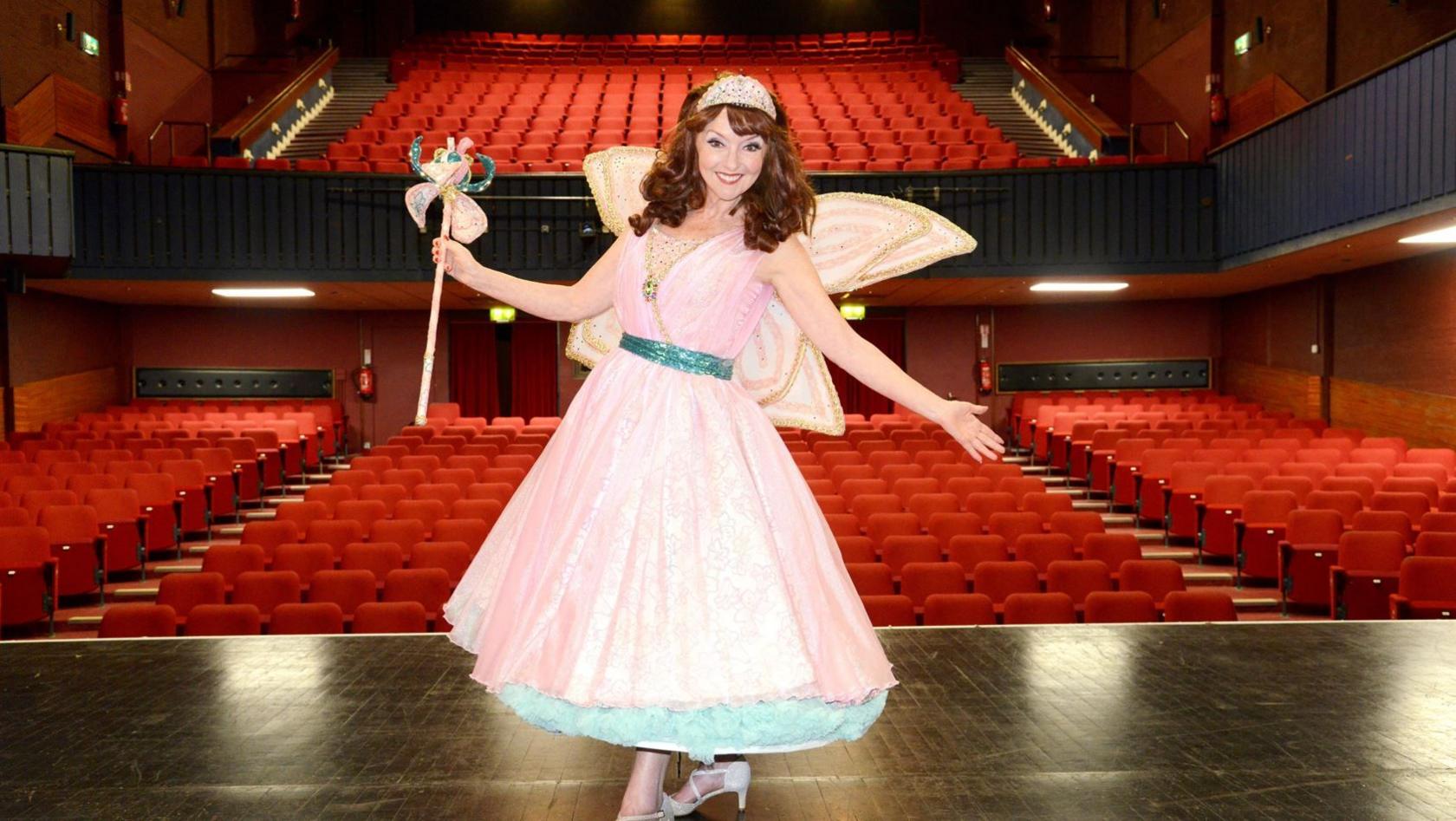 Melanie Walters standing on stage wearing a pink fairy dress with blue ribbon and petticoat. She is wearing glittery pink wings, a tiara, white heels and is holding a pink wand in the air. Behind her is a theatre with hundreds of red cushioned seats. 