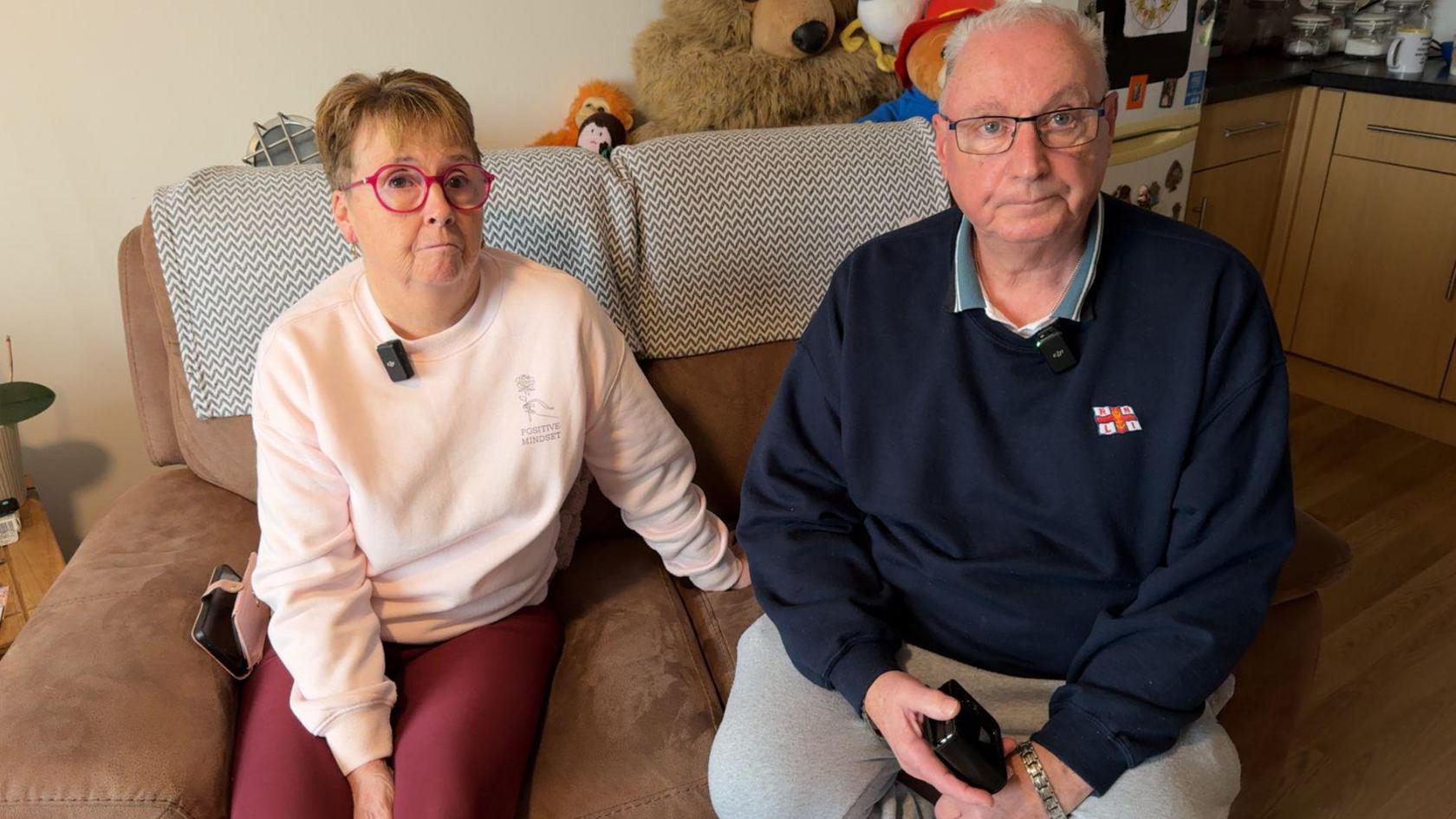 A woman with a pink jumper, pink glasses and short hair sitting on a brown sofa next to a man with short, white hair who is wearing a navy RNLI jumper and black glasses.