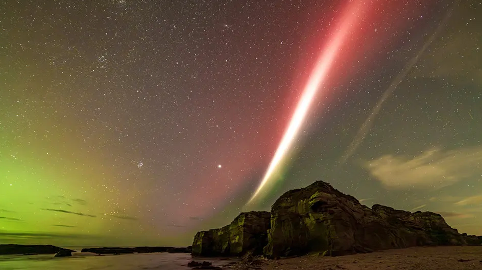 A large rock on a beach with the aurora in the sky above, and also a STEVE - a streak of very bright light with a tinge of red and green running through the sky