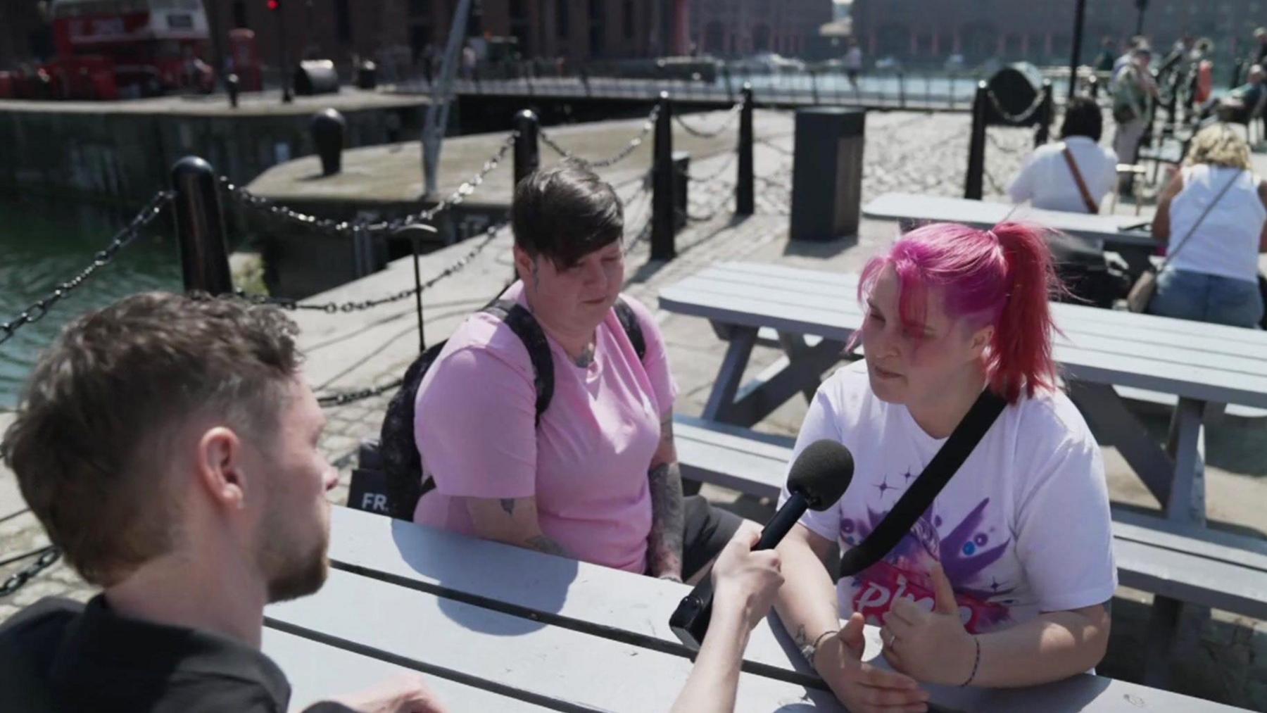 Newsbeat reporter Jordan Kenny, a male, holding a microphone and interviewing two women while sitting at a table 