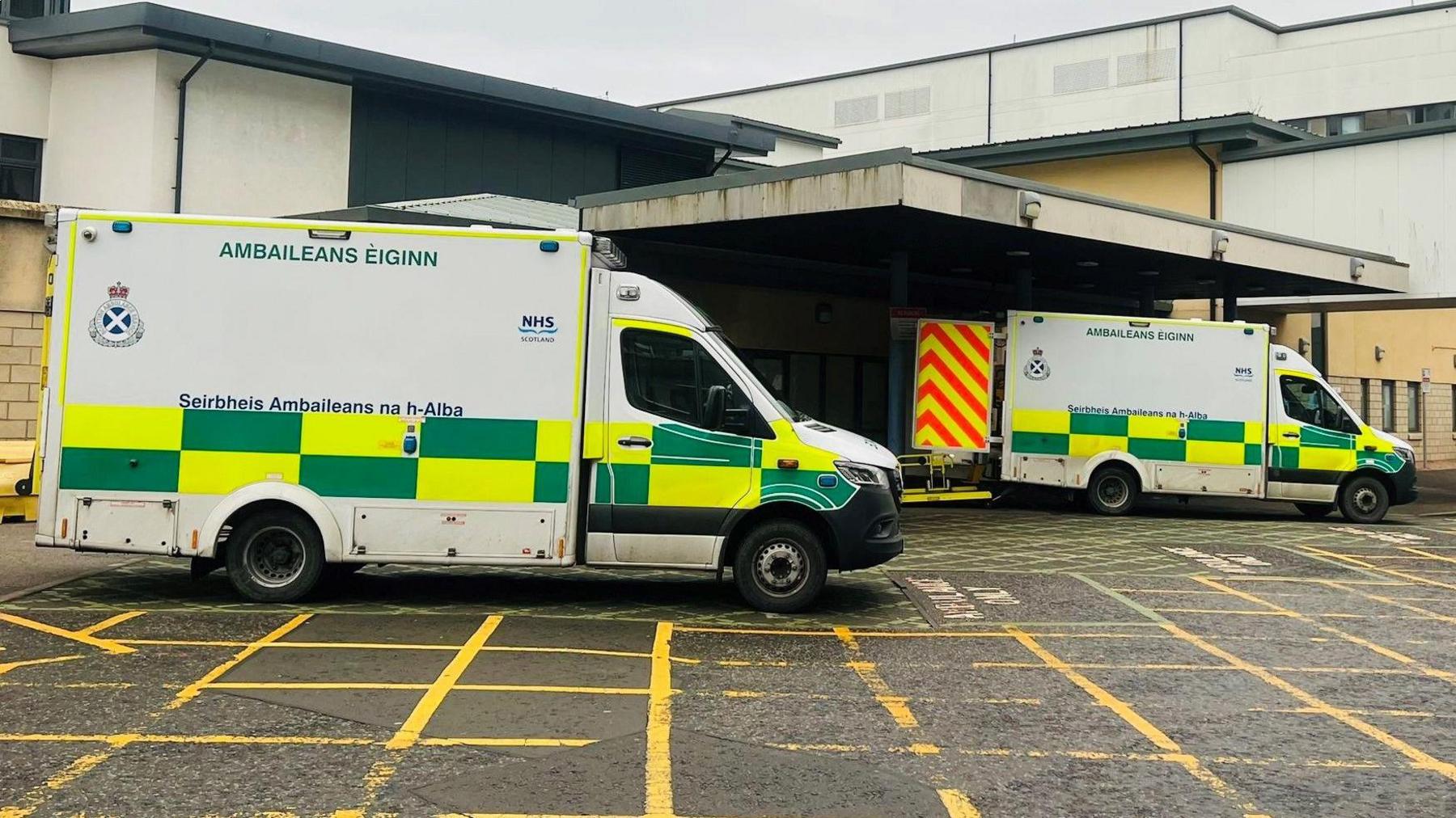 Ambulances with green and yellow checked markings, parked outside a hospital building.