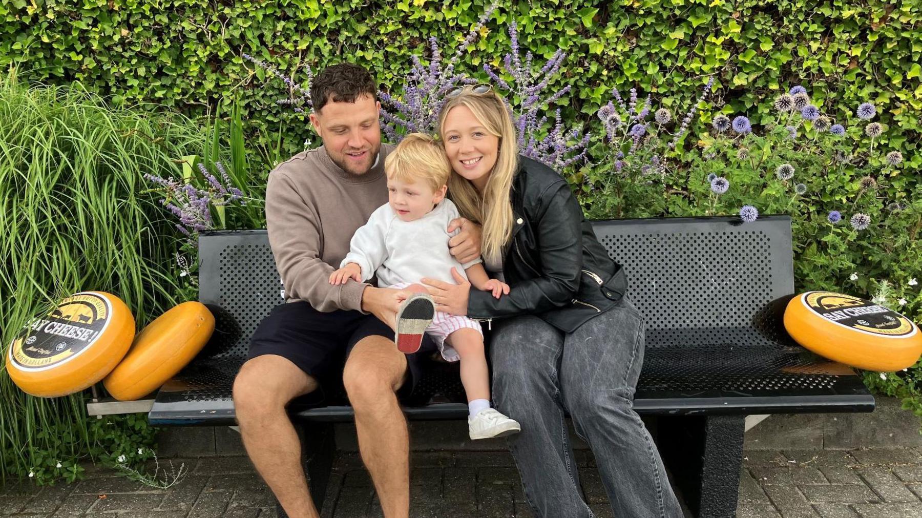 The couple sits on a black metal bench with big, yellow cheese on either side. They hold their son Archer between them in the middle.