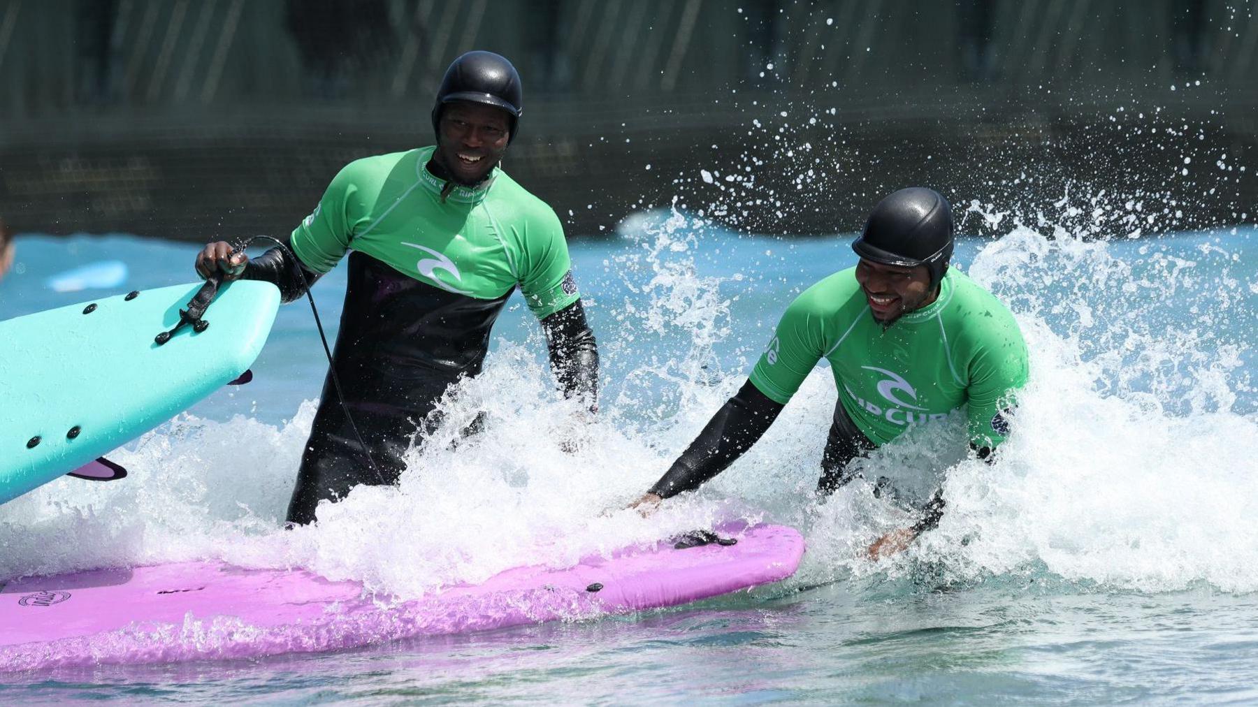 Two men stand in the water at The Wave in Bristol. They are smiling and wearing green and black wetsuits as the water foams around them