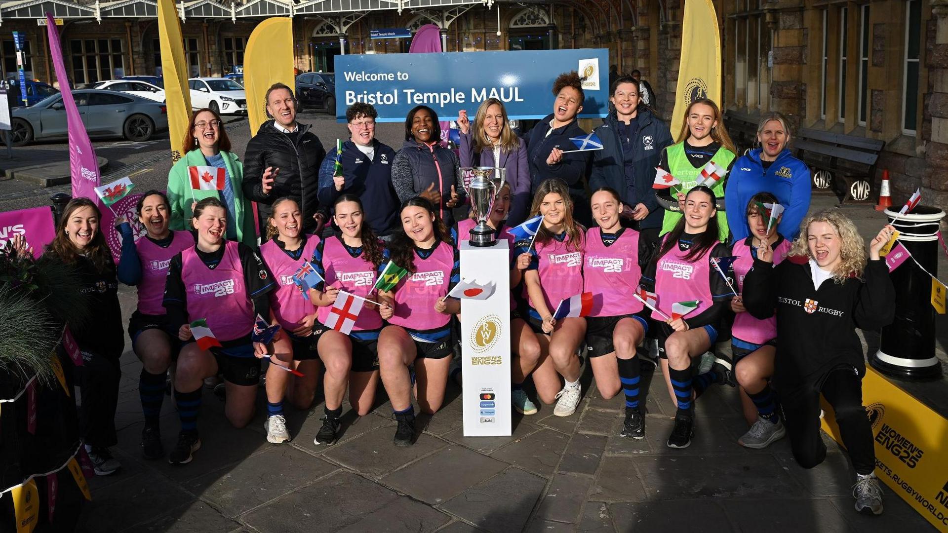 A large group of people stand outside Bristol Temple Meads station to launch ticket sales for the Women's Rugby World Cup 2025. Behind them is a large sign showing the name "Bristol Temple Maul" It is a sunny day and the group, mostly women, is smiling at the camera