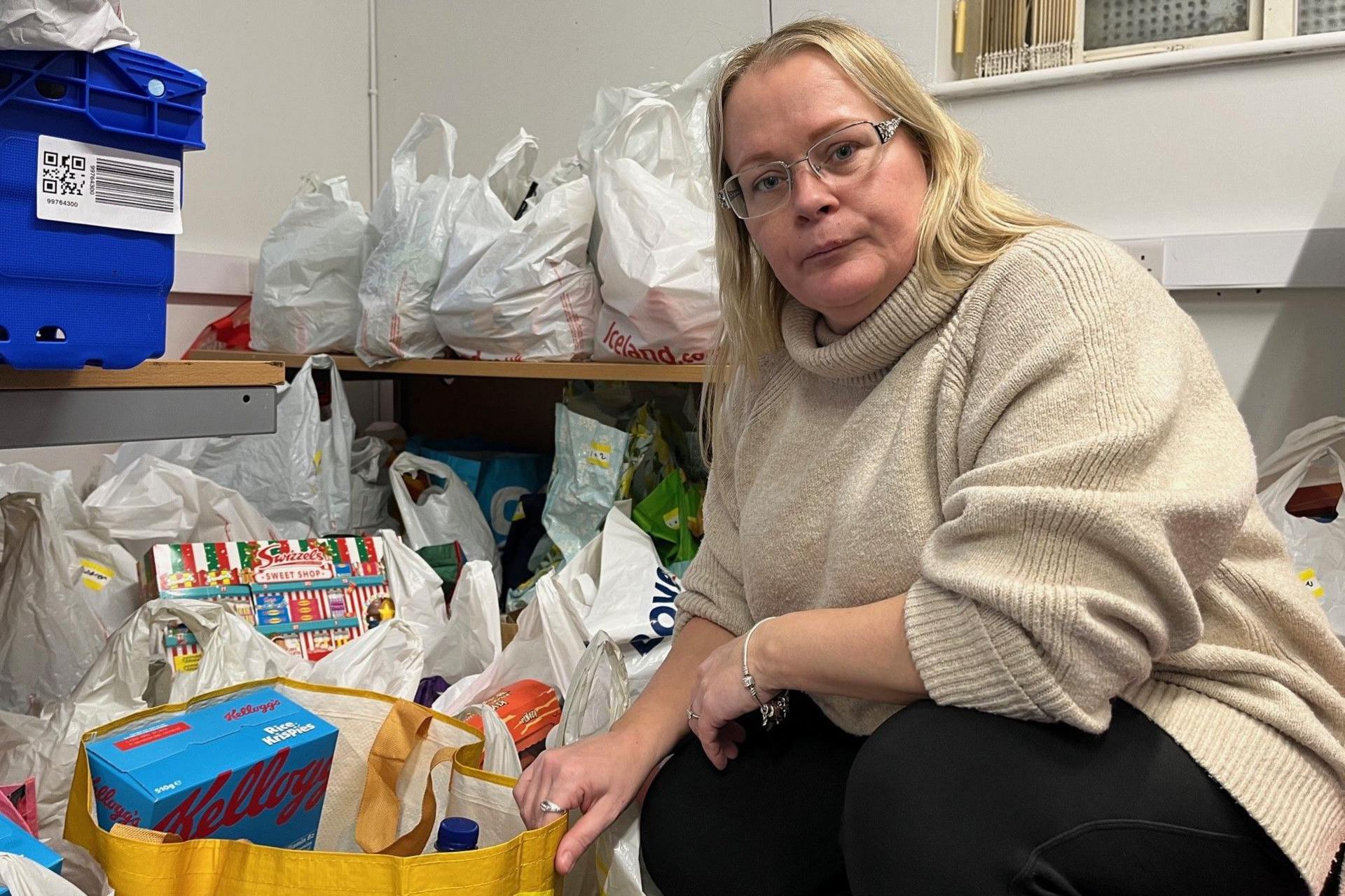 Samantha Palmer, a food bank co-ordinator, wearing a cream-coloured pullover and black trousers, crouches down next to dozens of carrier bags full of donated food