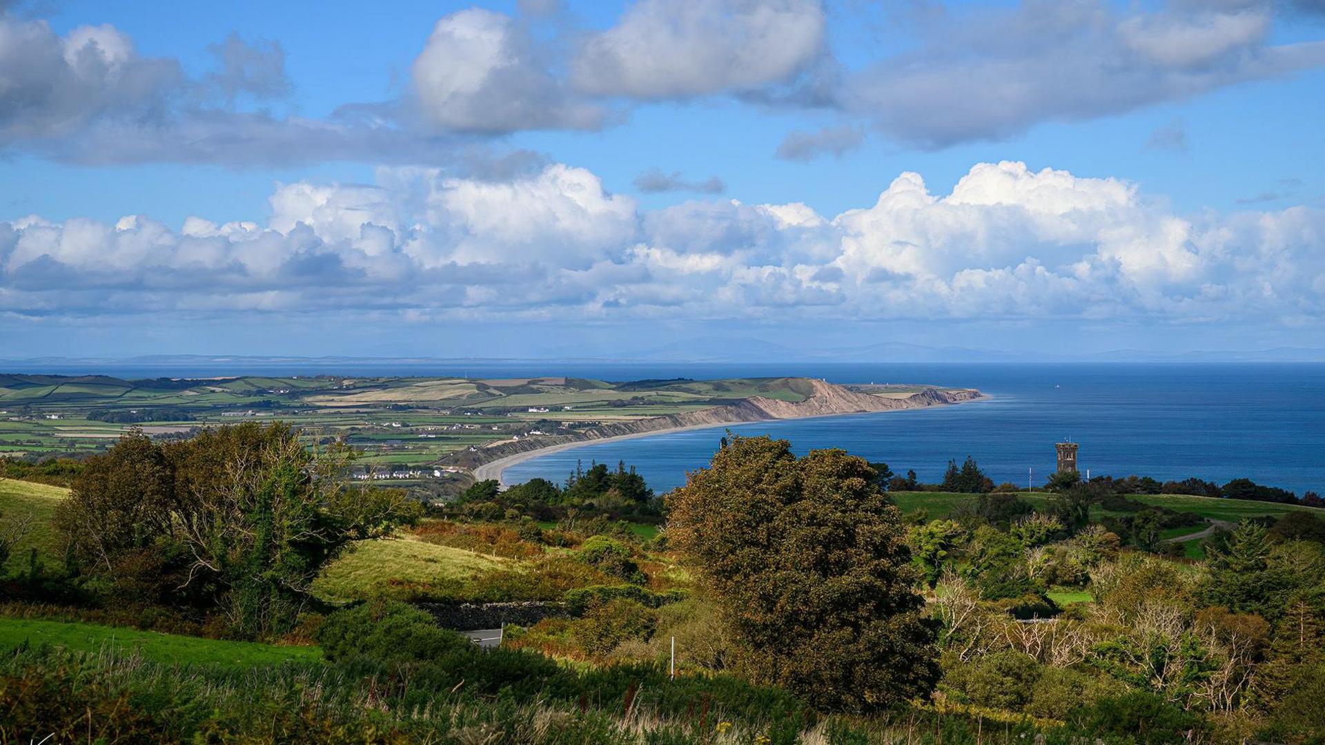 A view to the northern tip of the Isle of Man with a blue cloudy sky, blue sea, and a foreground of green hills dotted with trees. Houses can also be seen in a town that sits along a stretch of sandy beach. 