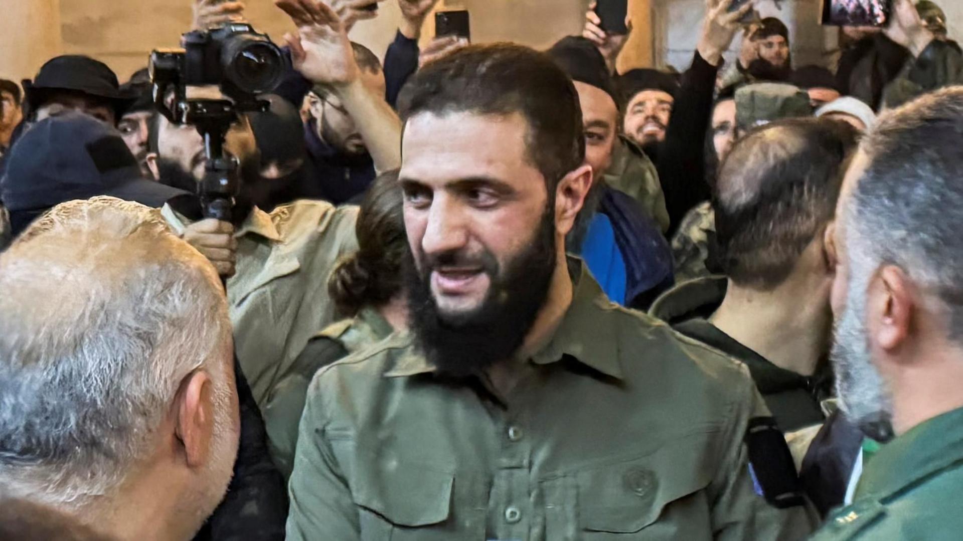 Ahmed al-Sharaa, previously known as Abu Mohammed al-Jolani, the leader of HTS, speaking to a group of supports inside the Ummayad Mosque in Damascus. Many people are taking photographs of him. He is wearing a green military-style shirt.