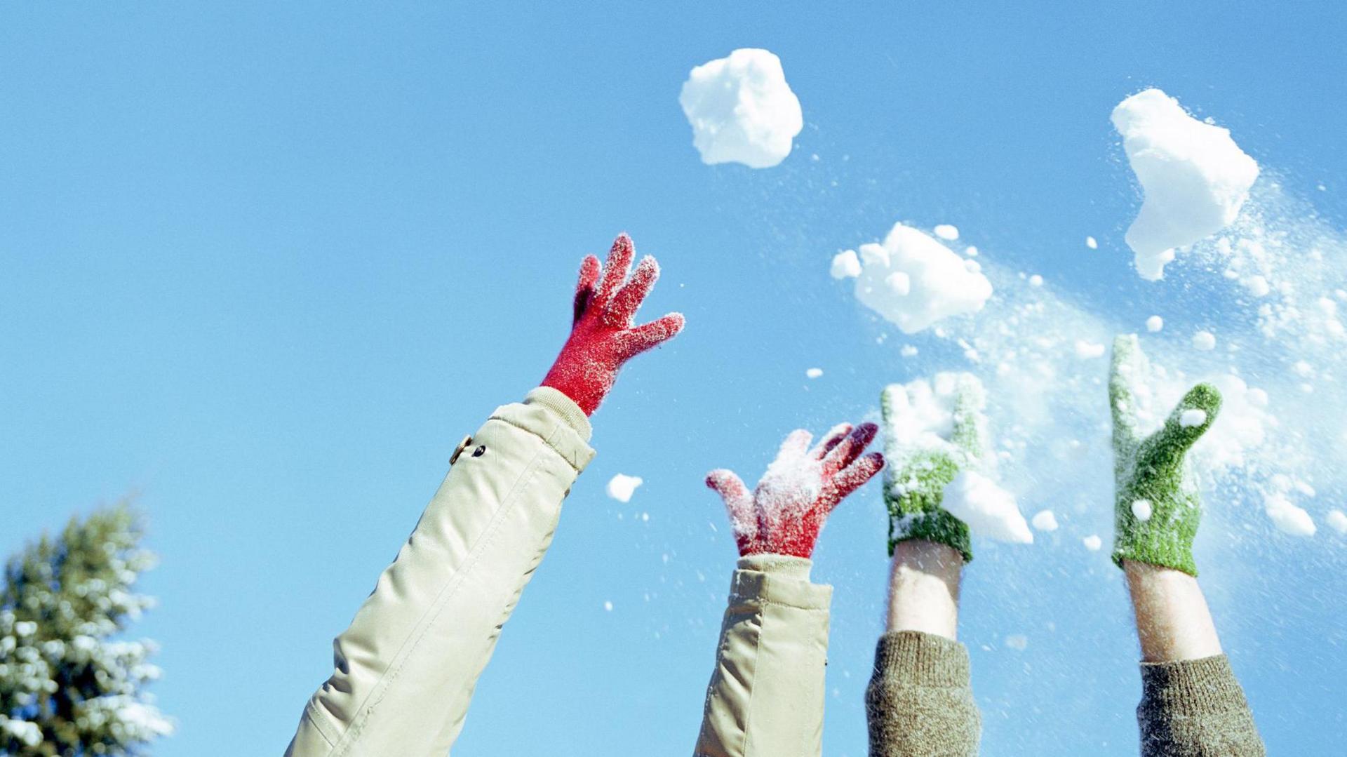 Close up of two sets of arms and hands (one pair in red gloves and another in green ones) throwing snowballs