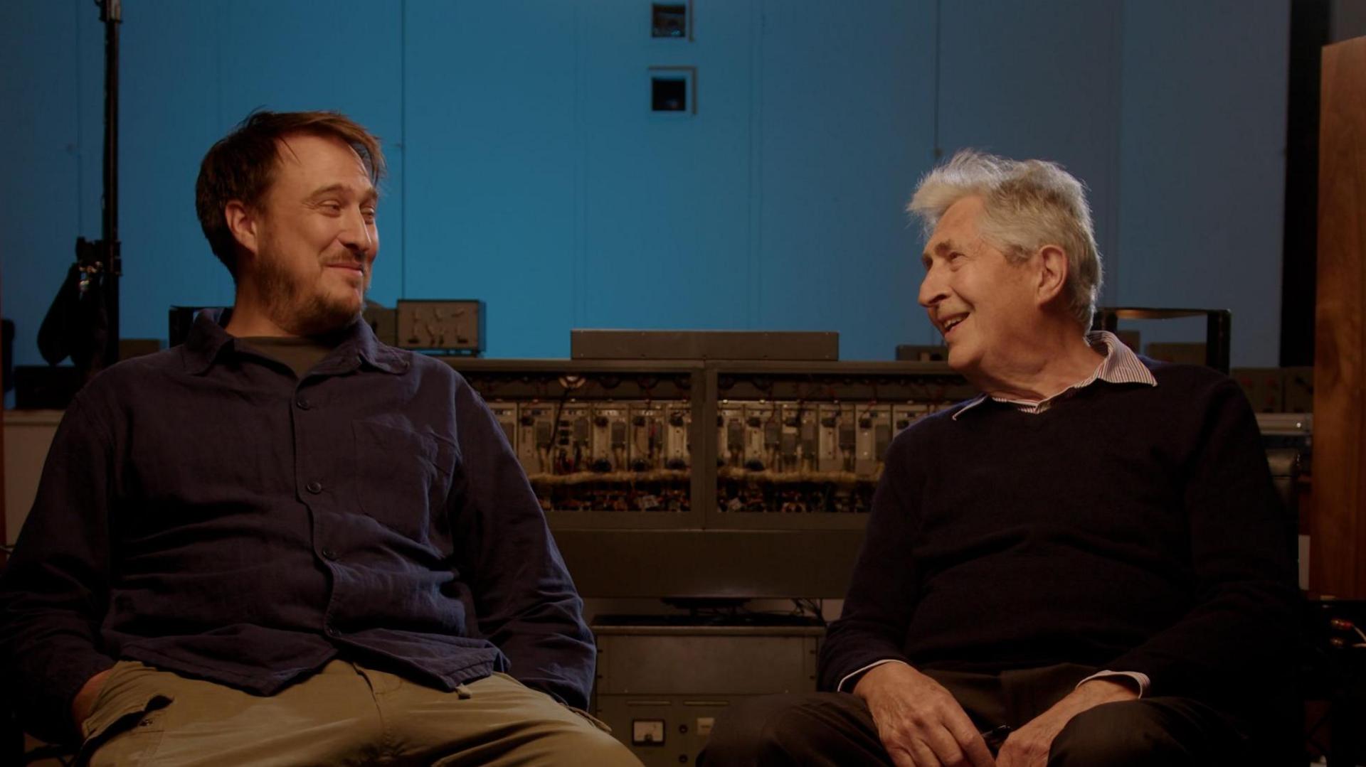 Hamish and Malcolm sit beside each other smiling at one another. They are sitting in a music studio with sound equipment behind them as well as a recording booth. Hamish has dark short hair, is wearing a blue shirt and brown trousers. Malcolm has grey short hair and is wearing a black jumper with a light shirt on underneath.