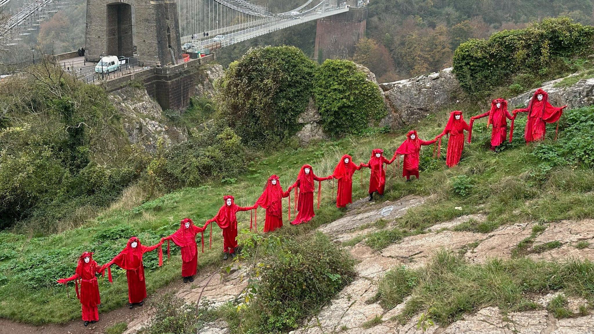 A line of women dressed from head to toe in red stand with the Clifton Suspension Bridge in the background. The group, known as the Red Rebel Brigade, appeared in various Bristol locations to highlight the start of COP 29 in Baku