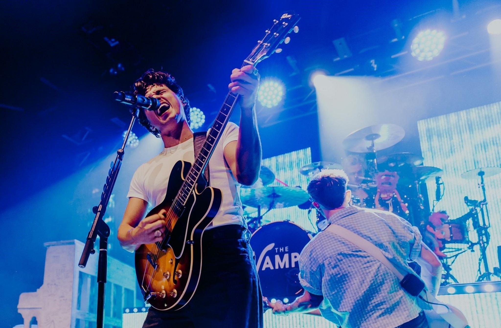 Brad Simpson of The Vamps performs at O2 Academy Bristol. His eyes are closed as he sings passionately into the microphone, while also playing his electric guitar. He is wearing a white T-shirt and dark trousers, and other members of the band can be seen in the background