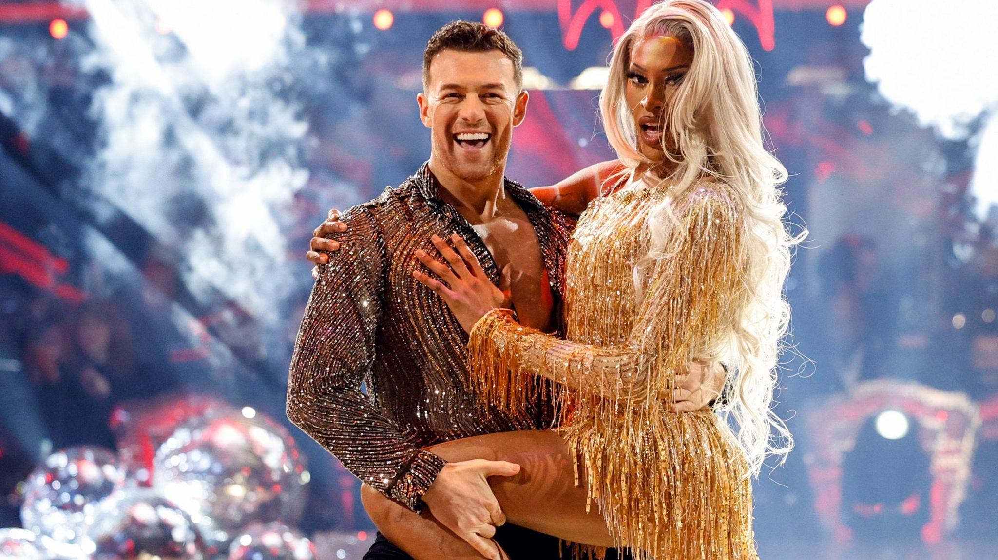 Tayce dancing with Kai Widdrington on Strictly. Tayce is wearing a short gold fitted dress and has long blonde hair. Kai is wearing a sheer black and silver shirt and black trousers and there is a Christmas tree in the background