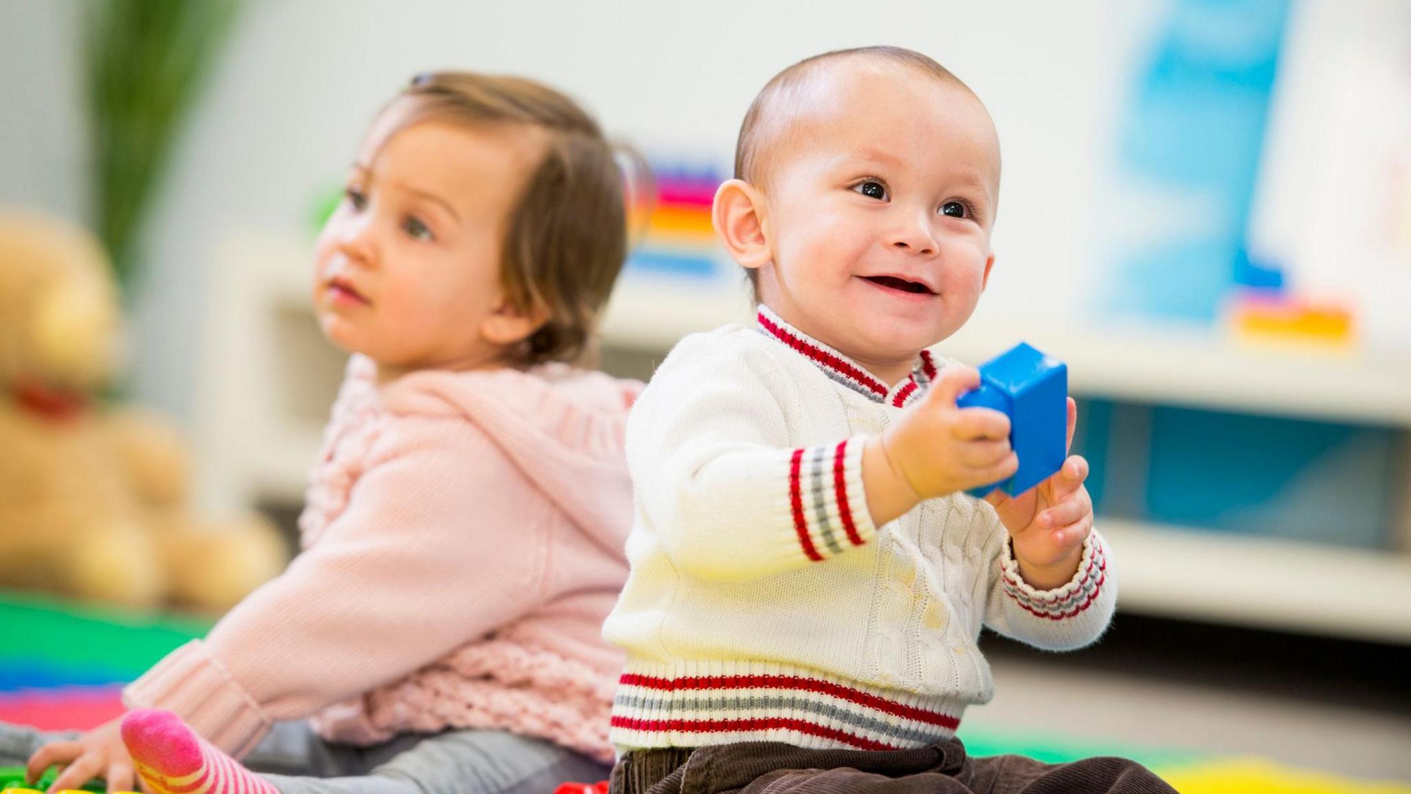 Two babies sit on the floor of a playroom dressed in jumpers, one looking ahead and holding a toy, and the other in the background looking off into the distance 