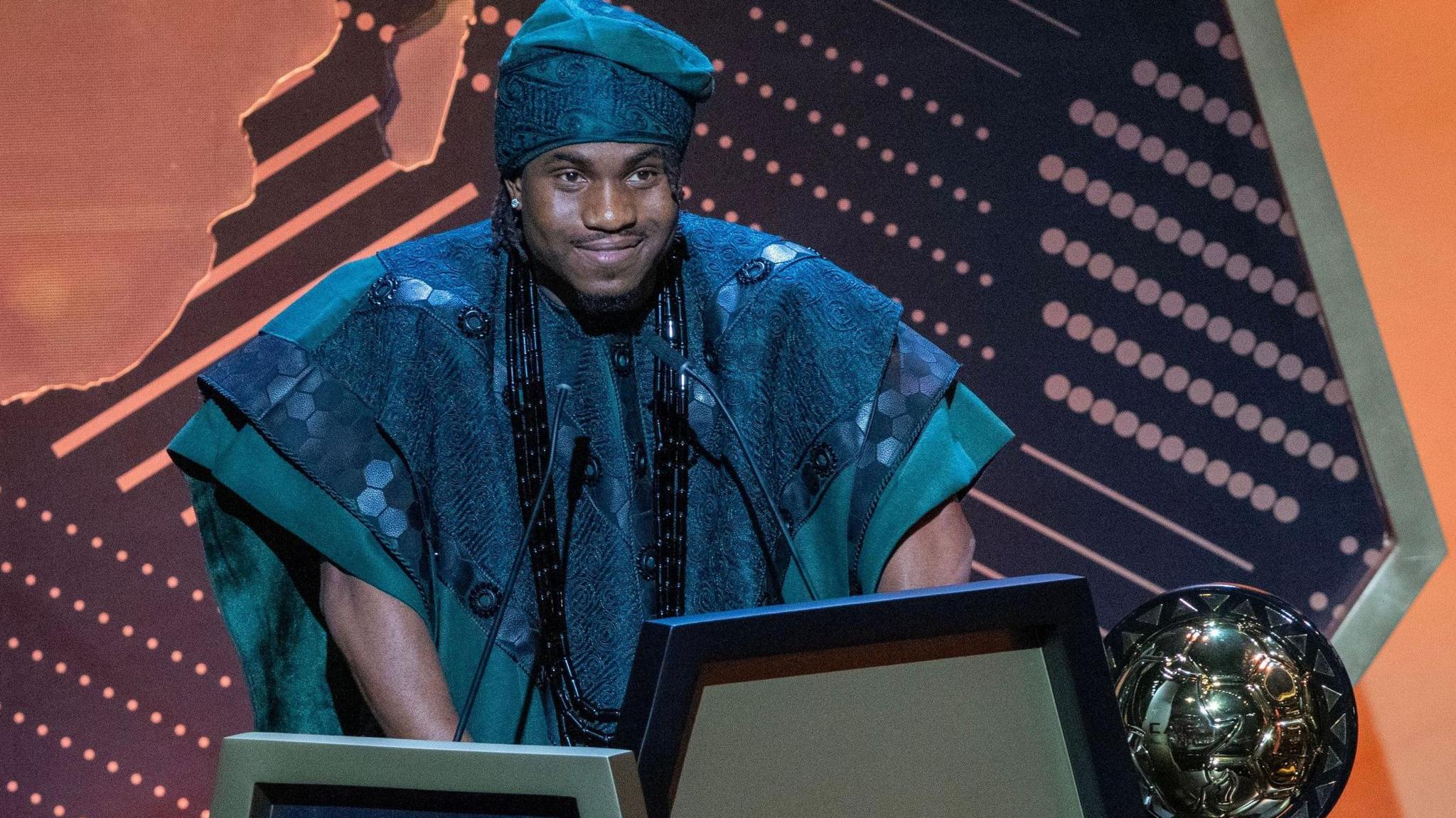 Ademola Lookman, wearing a traditional dark green Nigerian robe and matching hat, stands at a podium to give a speech after being named African Footballer of the Year