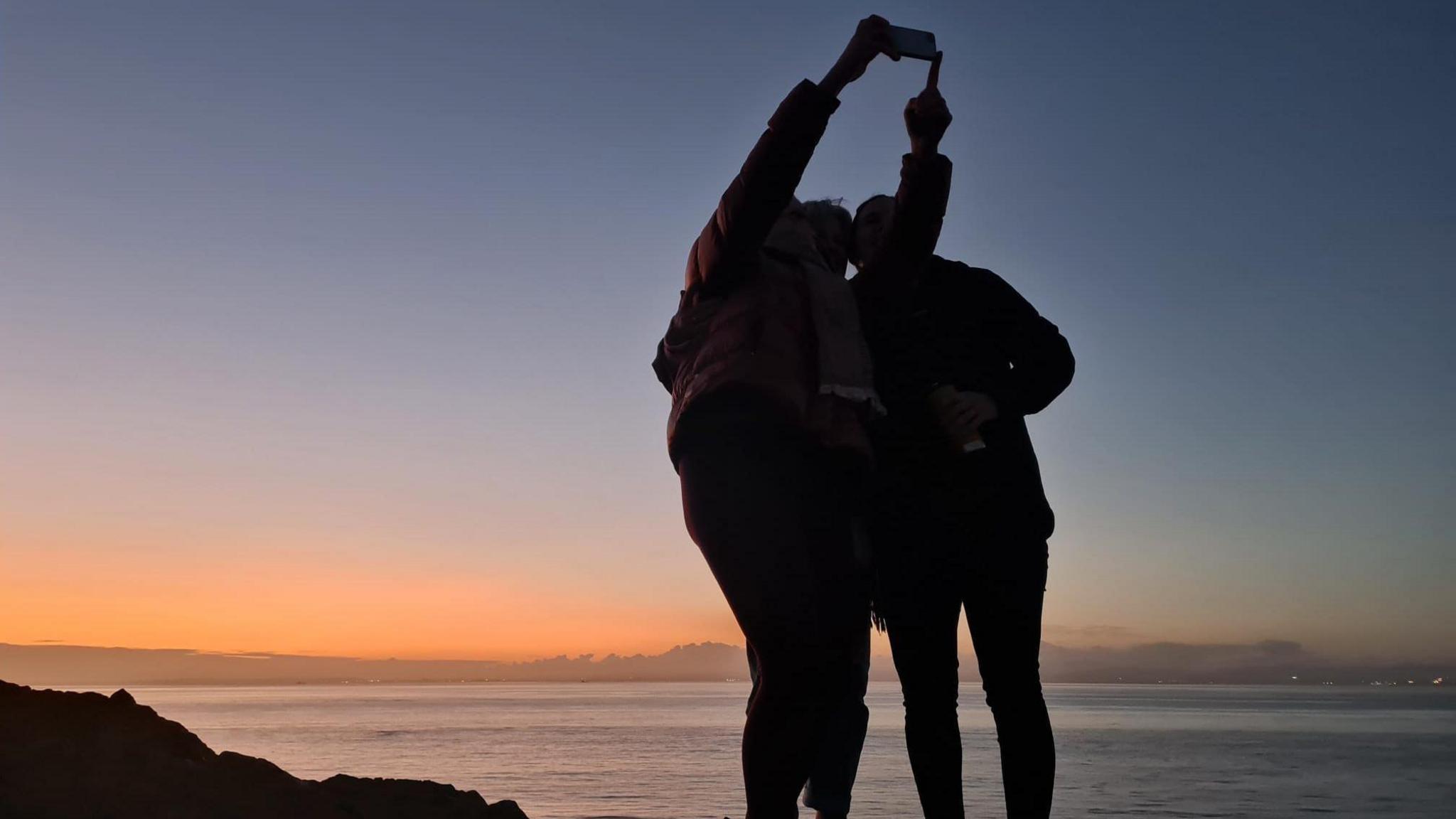 Three women are silhouetted as they pose for a selfie on the Somerset coast at Clevedon. Behind them the setting sun has cast a light orange glow over the Severn Estuary