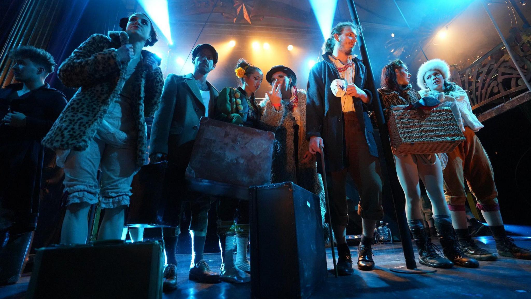 A group of Invisible Circus performers stand on stage in Bristol. They are in various vintage costumes and are lit by blue lights