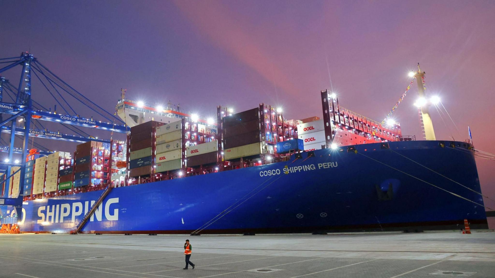 A man walks at China's state-owned Cosco Shipping Chancay port inaugurated during the APEC Summit, in Chancay, Peru. There is a large container ship framed by a colourful sunset. 