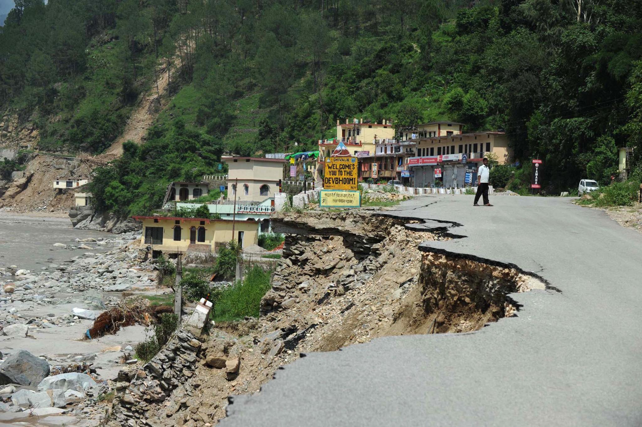 A road beside a river has been washed away by flood water in India.  A small town can be seen in the background.