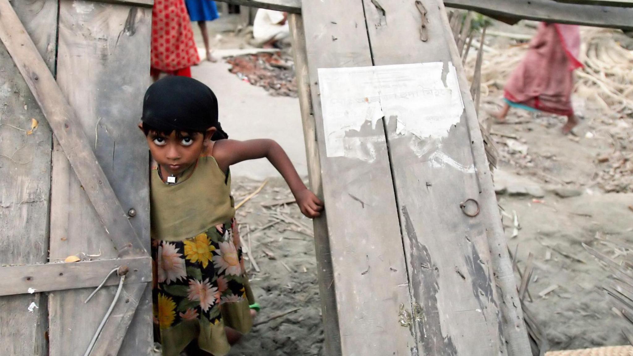 A young girl with dark hair and big eyes looks out through the doorway of a wooden building that has been shattered by the storm.  She looks sad and anxious.