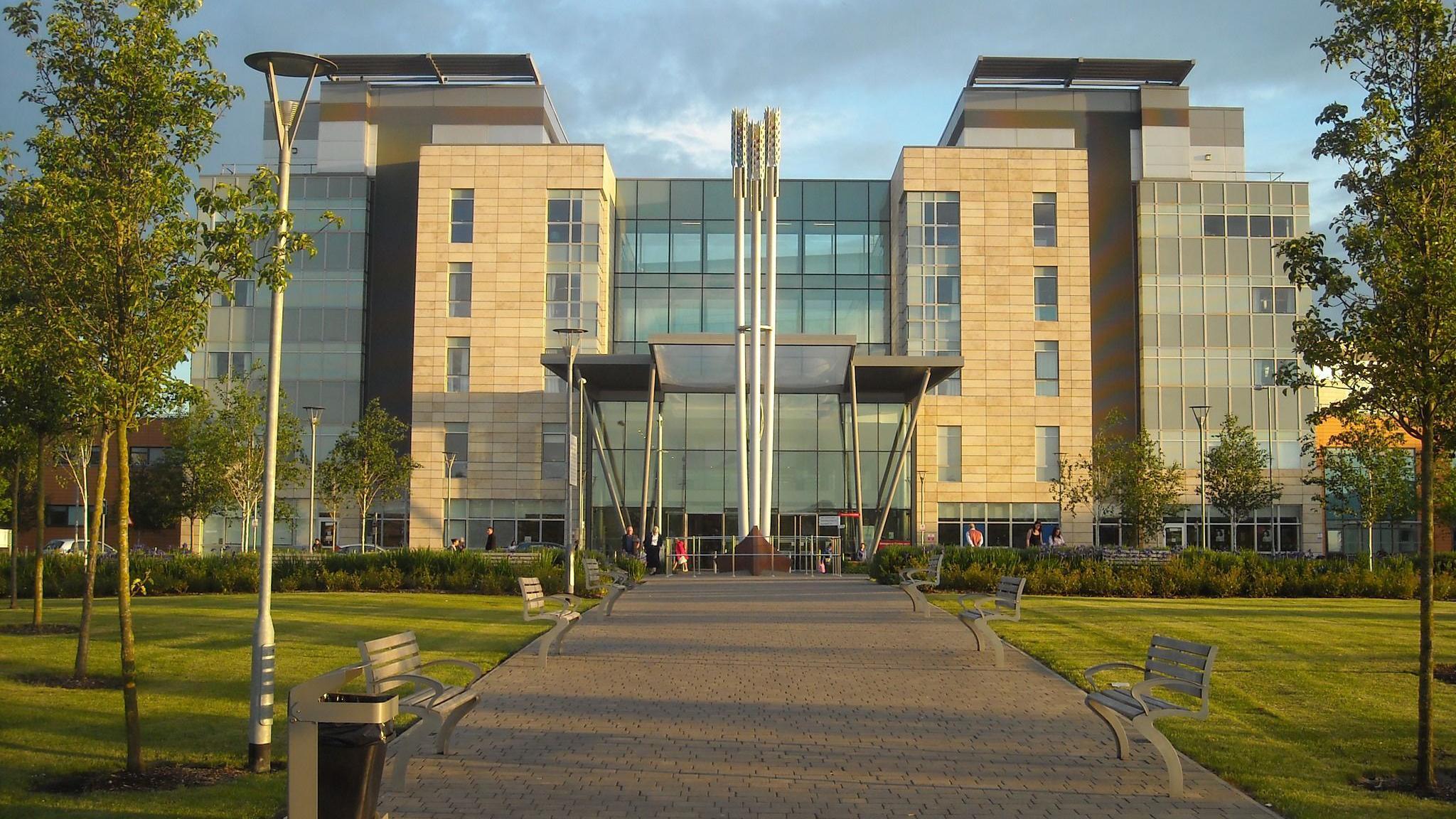 The computer-generated image of the exterior of Peterborough City Hospital. A large glass-panelled building with a central walkway with green space on either side. The walkway has benches and is lined with trees.
