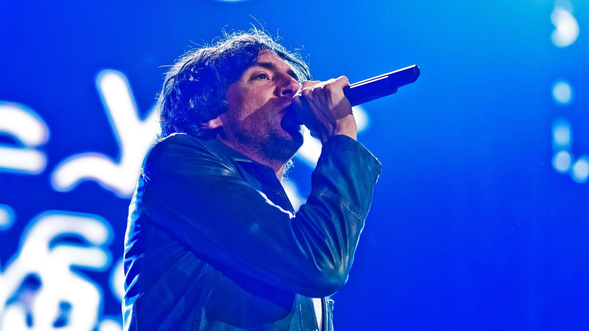 Snow Patrol singer Gary Lightbody sings into a microphone on stage wearing a blue jacket. Behind him is a blue screen with white letters.