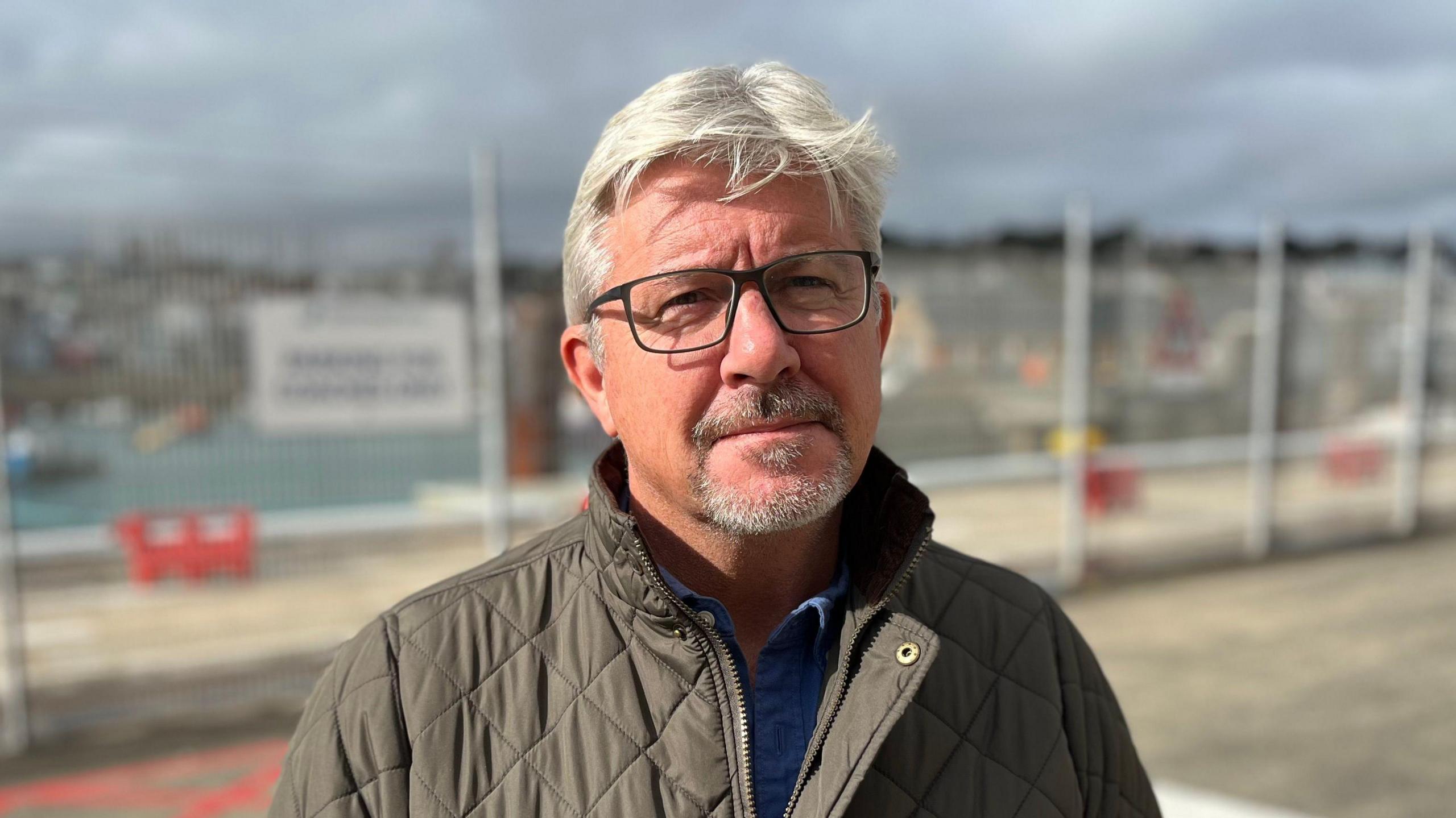 Deputy Helyar stood in St Peter Port. the background is blurred. He is wearing a grey jacket and a blue shirt. He has short grey hair and a grey goatee beard. He is wearing dark framed glasses.