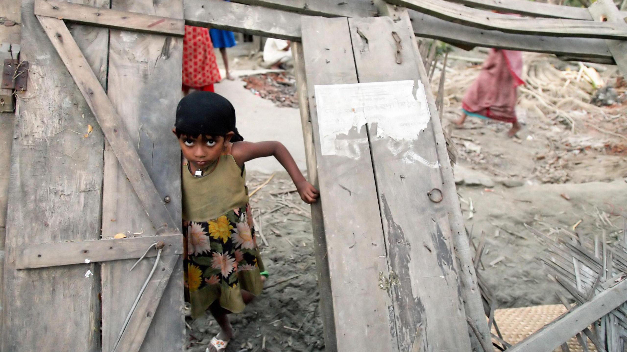 A young Bangladeshi girl wearing a green dress with flowers on the skirt stands in the wooden doorway of her collapsed house. 