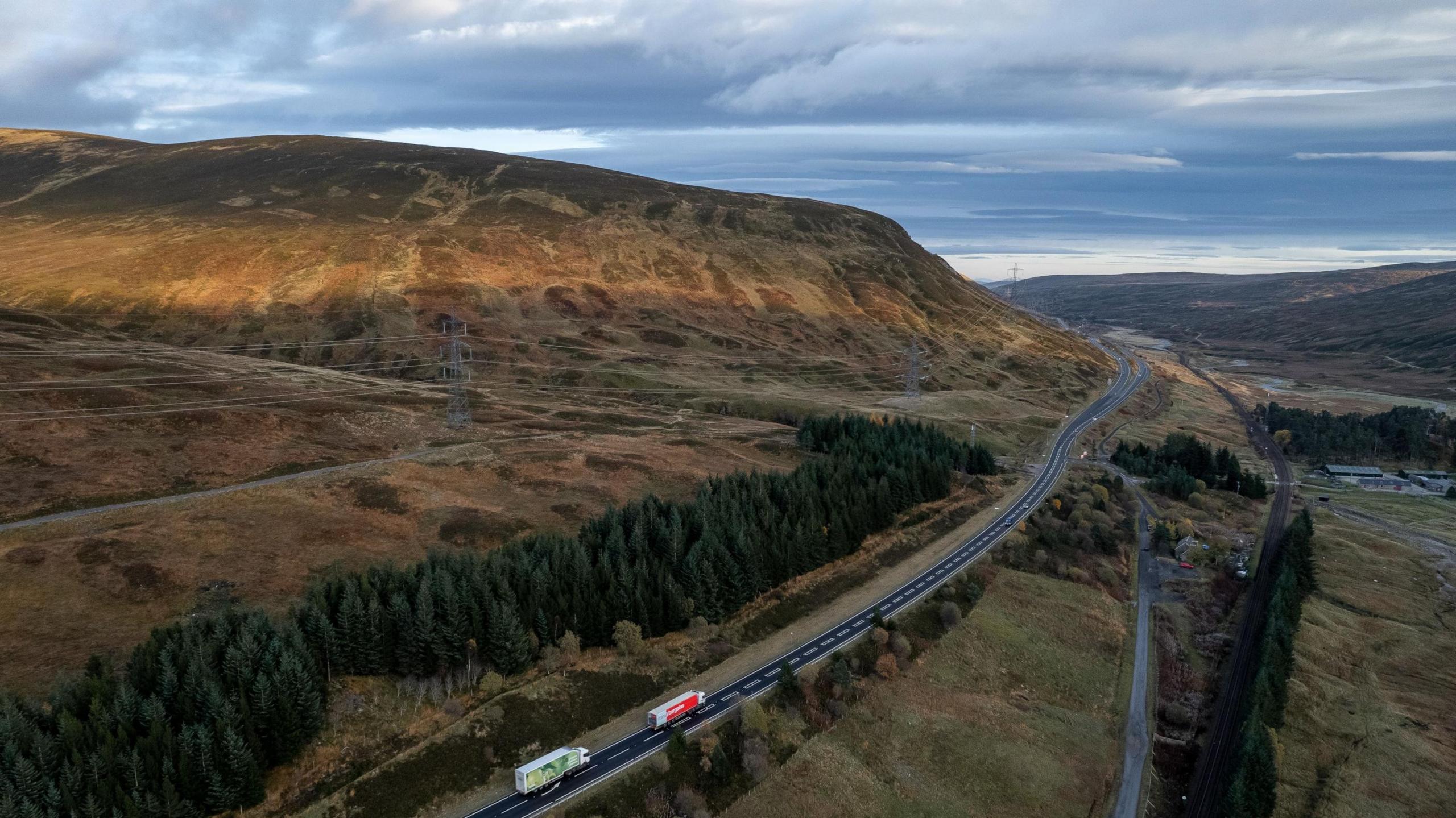 An aerial view of the A9 at Glen Garry. There are two lorries, both white, one with a green trailer and the other with a red trailer, on the road in the foreground. In the background, greenery including trees and grass can be seen going up the side of a hill, which is partially in the sun. The sky is blue and grey.