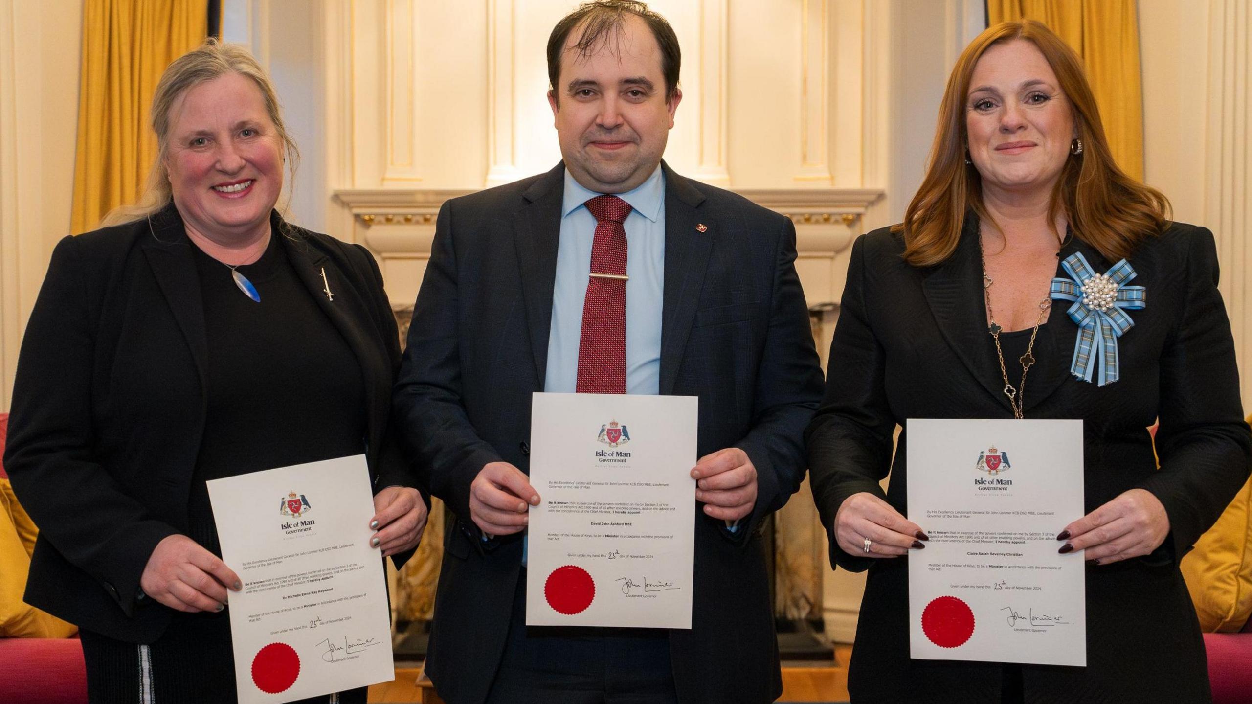 Michelle Haywood, who is wearing a black suit and has fair hair, David Ashford, who is wearing a dark blue suit, blue short and red striped tie and has dark hair, and Claire Christian, who is wearing a black suit with a blue Manx Tartan bow brooch. They standing shoulder to shoulder holding up their certificates of office.