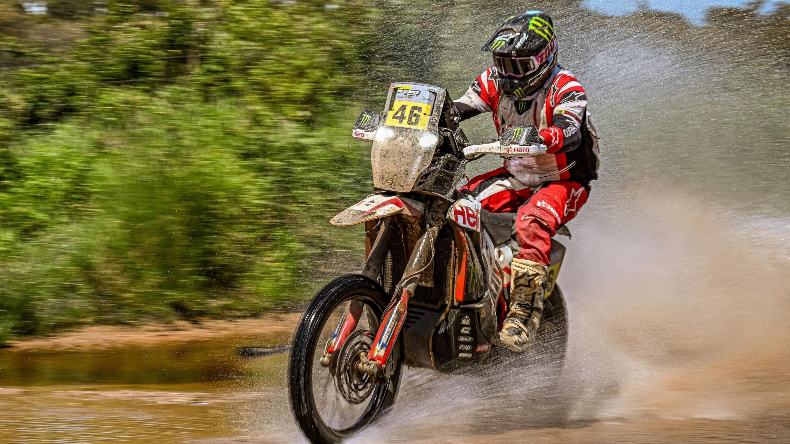 Ross Branch, wearing a black helmet and white, red and black overalls, rides a motorbike bearing the number 46 on the front through water which is spraying up beside and behind him while green foliage flashes past in a blur in the background