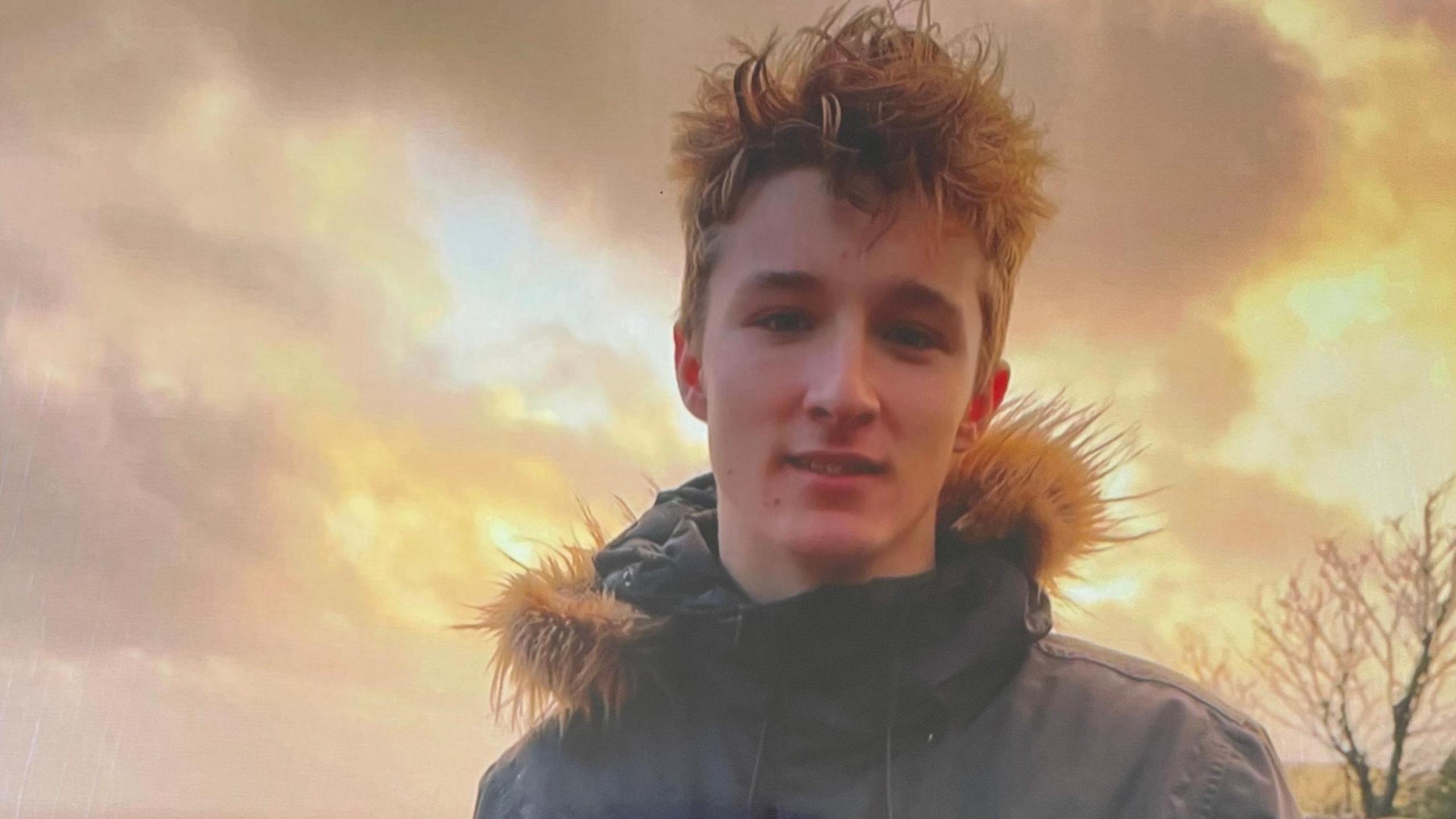 James, who has blond hair and is wearing a parka, looks at the camera. There is a tree in the background and he is set against a cloudy, orange sky.