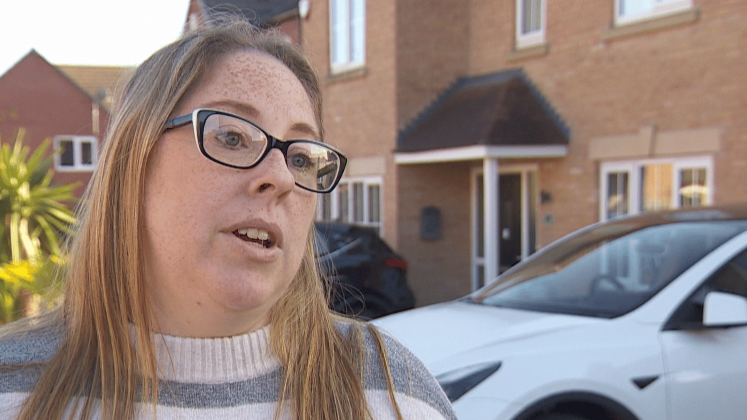 Charlotte Kelly stands in her new-build residential street in front of a brick-built house and a white car. She has long brown hair and wears black-framed glasses and a grey and white striped jumper.