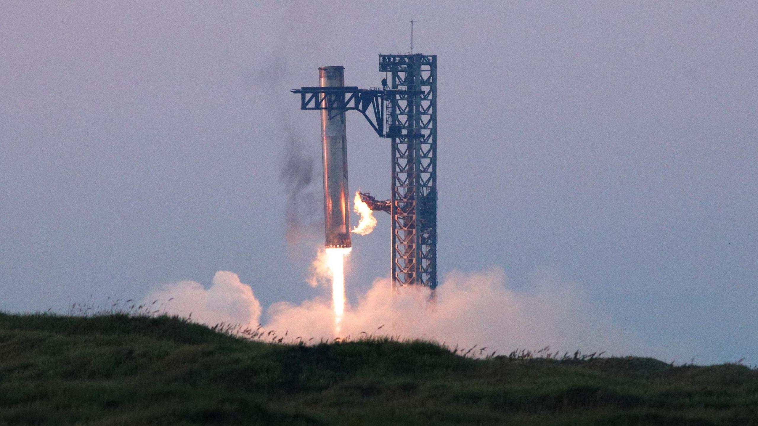 Starship's booster lowers itself into its launch tower as its jets power down. Fire is seen coming out of the bottom of the booster and smoke fills the surrounding air.