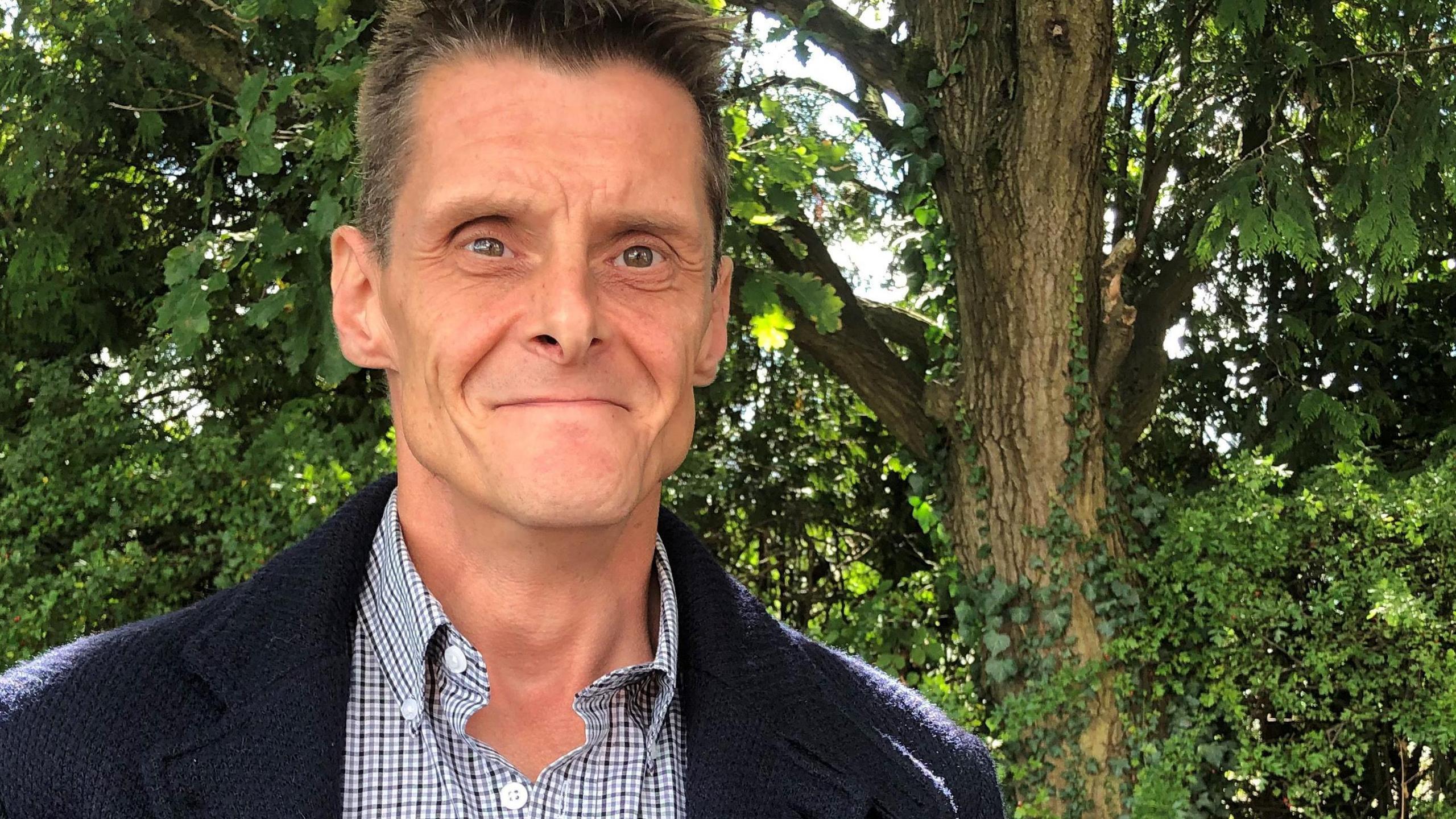 Jay Hornsby smiles at the camera as he stands with trees in the background, he is wearing a blue cardigan and chequered shirt. 