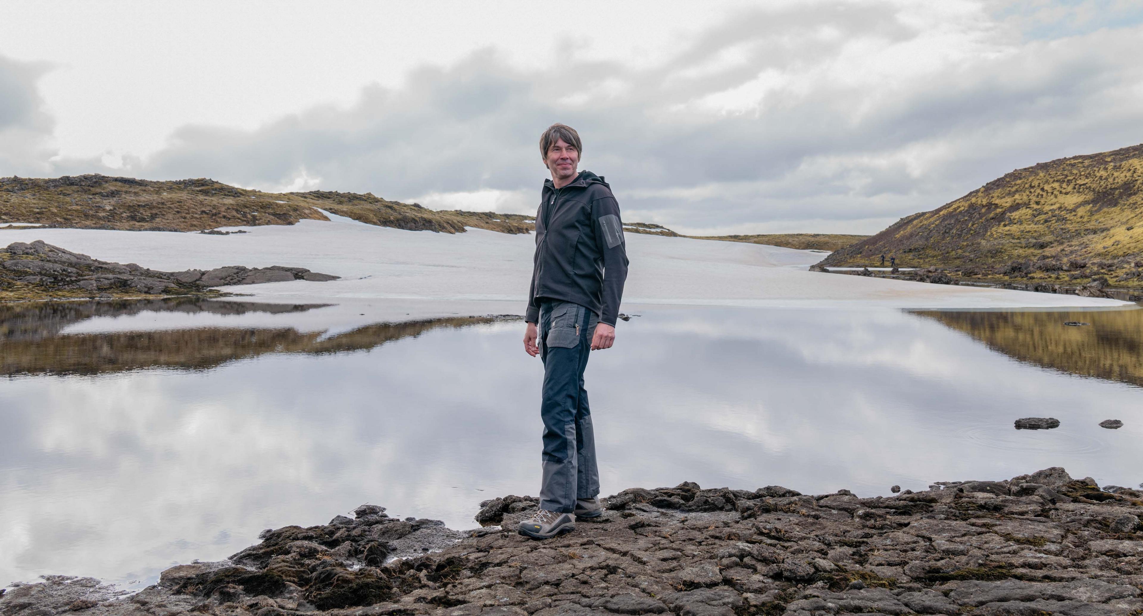 Prof Cox stands on some rocks at the edge of a large lake. Hills can be seen in the distance. He is wearing a grey hoodie, walking trousers and boots, and is smiling.