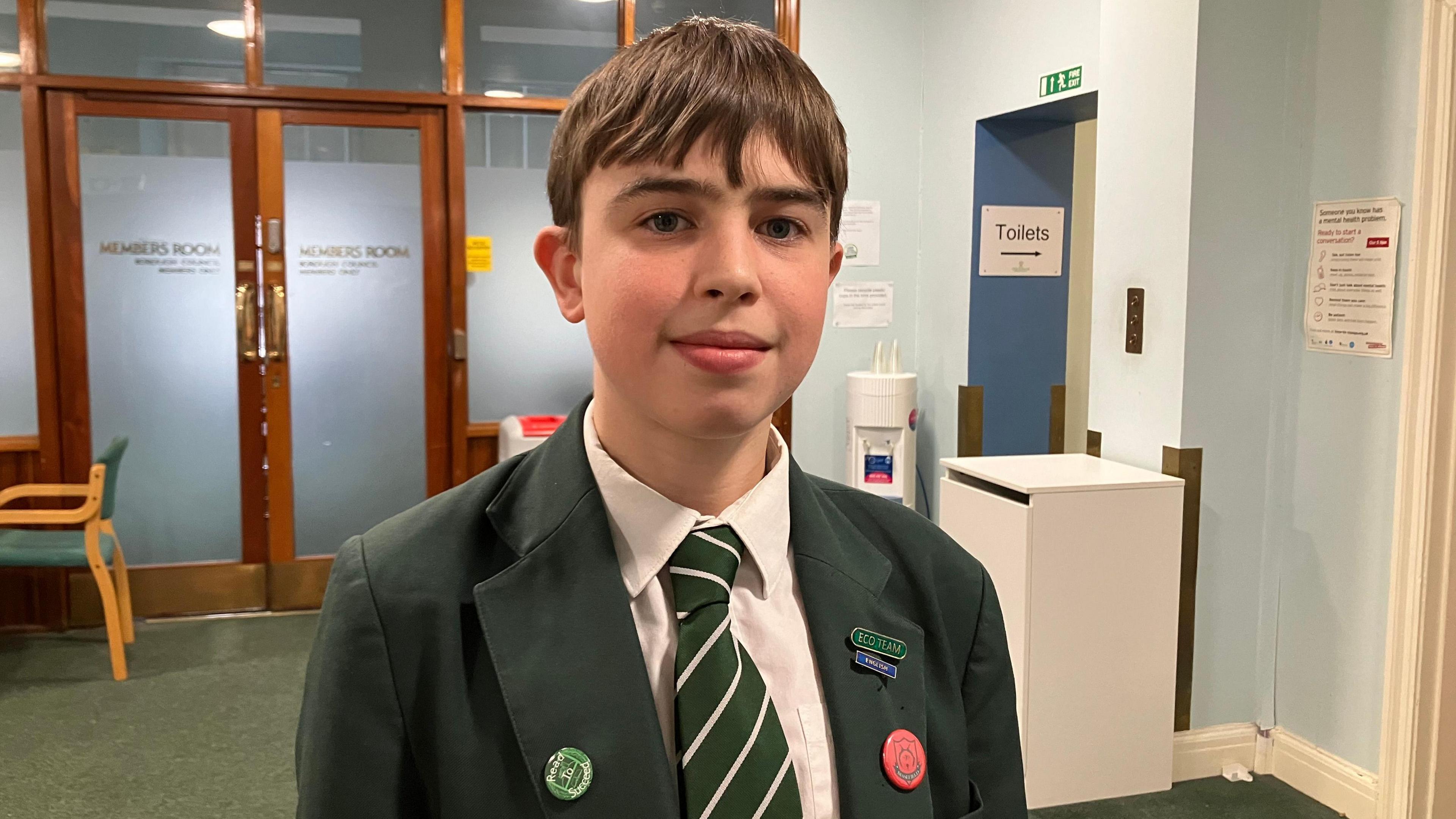 Leo, a 13-year-old boy with short brown hair, looks at the camera in a waiting room. He is wearing a dark blazer and a green and white stripey tie. There are enamel badges on his lapels.